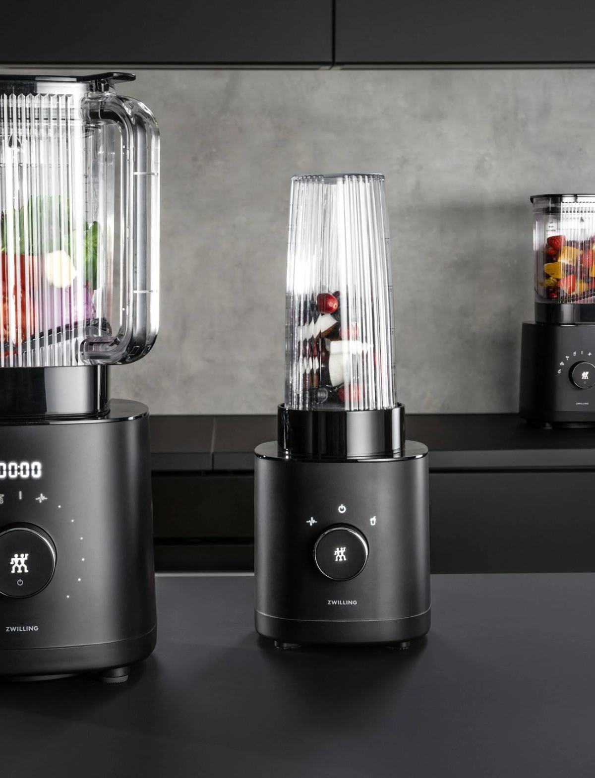 Drei schwarze Mixer mit Glasbehältern, einer mit Früchten, auf dunkler Oberfläche.
