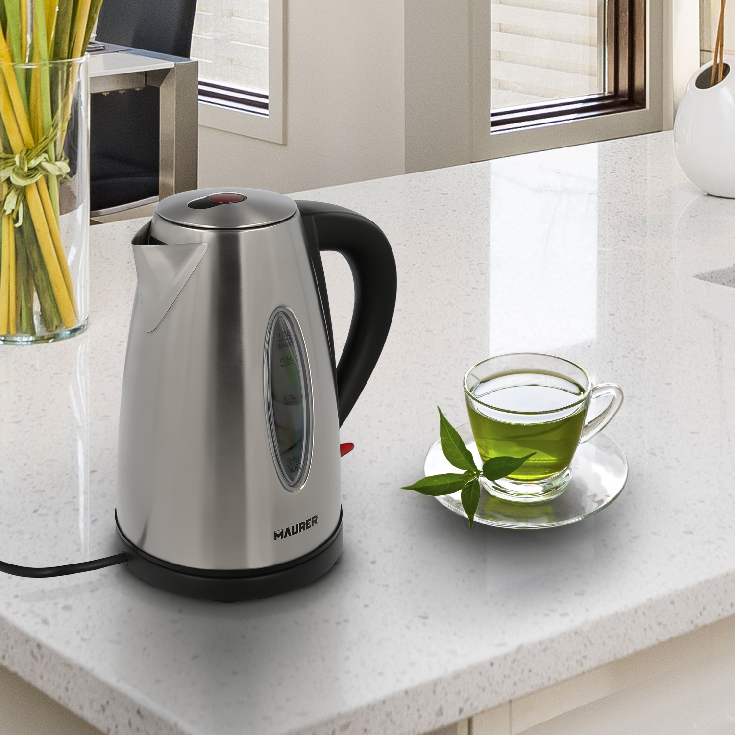 Una tetera de acero inoxidable con asa negra, un vaso de té verde y una hoja sobre una encimera.