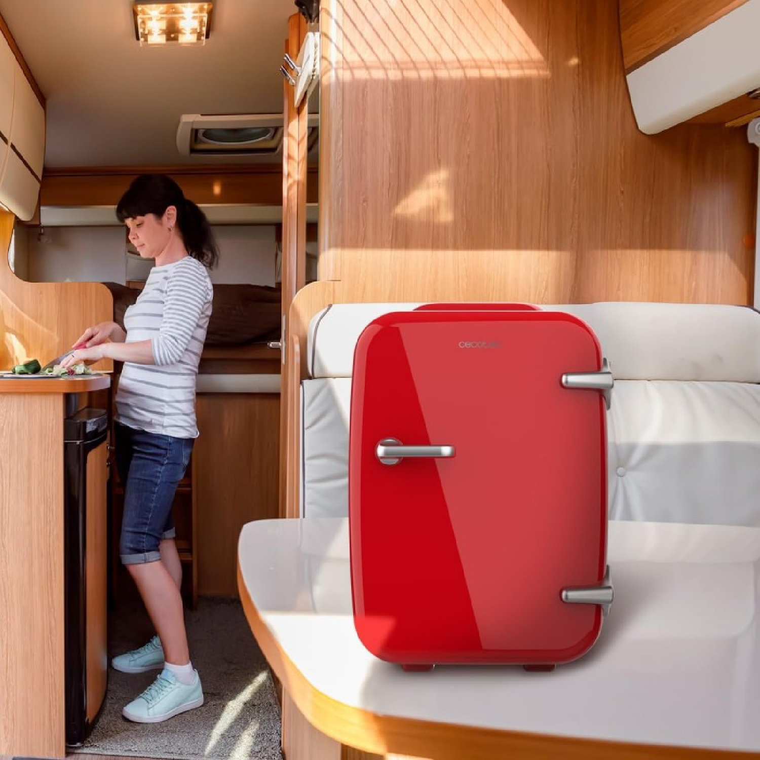 Mujer en cocina de RV, cortando verduras, nevera roja. Interior de madera, luz solar brillante, diseño moderno.