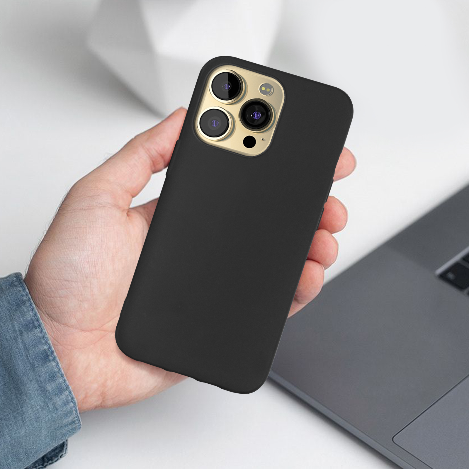 Een zwart telefoonhoesje met een gouden cameraring, vastgehouden door een hand. Een laptop is zichtbaar op de achtergrond.