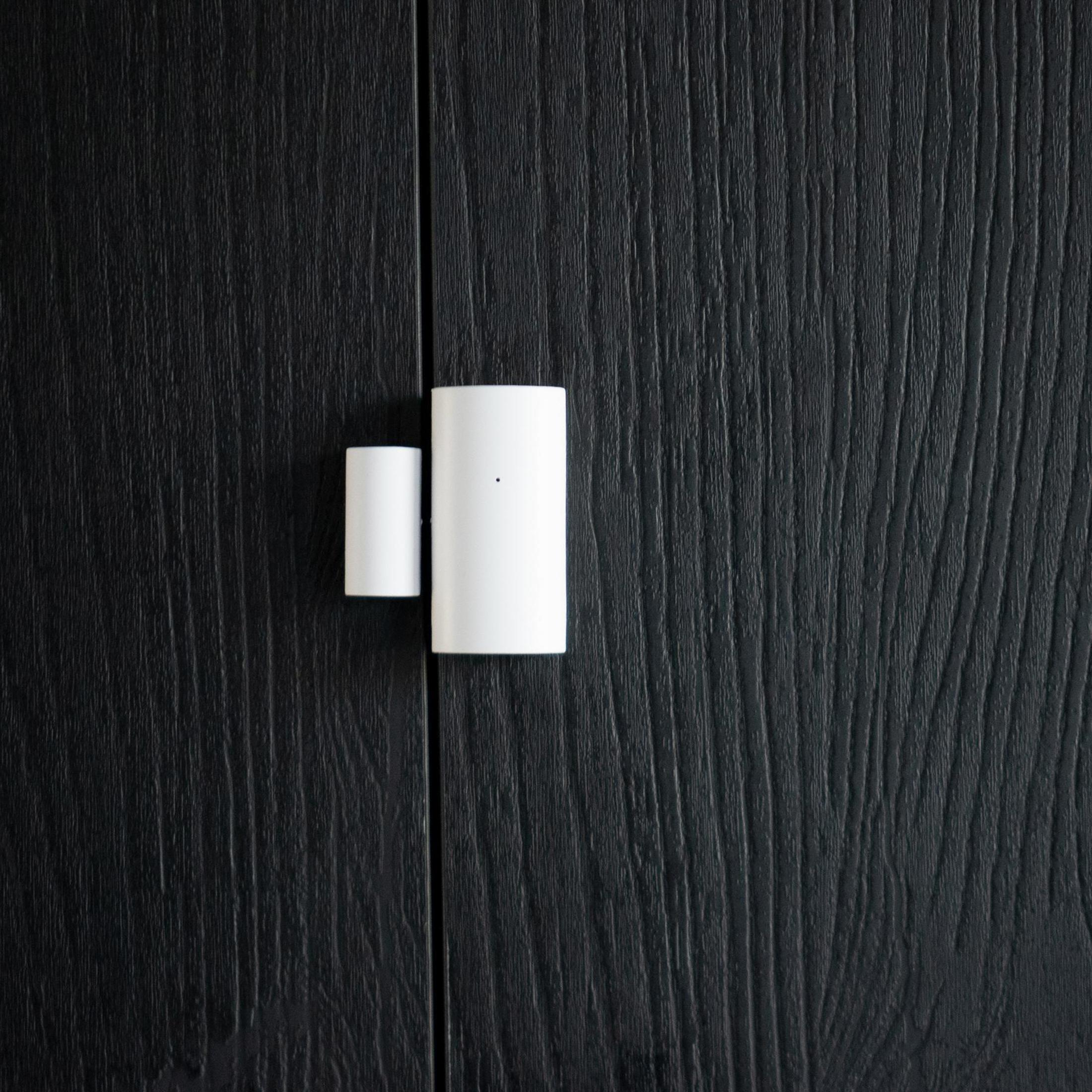 Weißer Smart-Home-Sensor an schwarzer Holztür, mit sichtbarem Spalt.