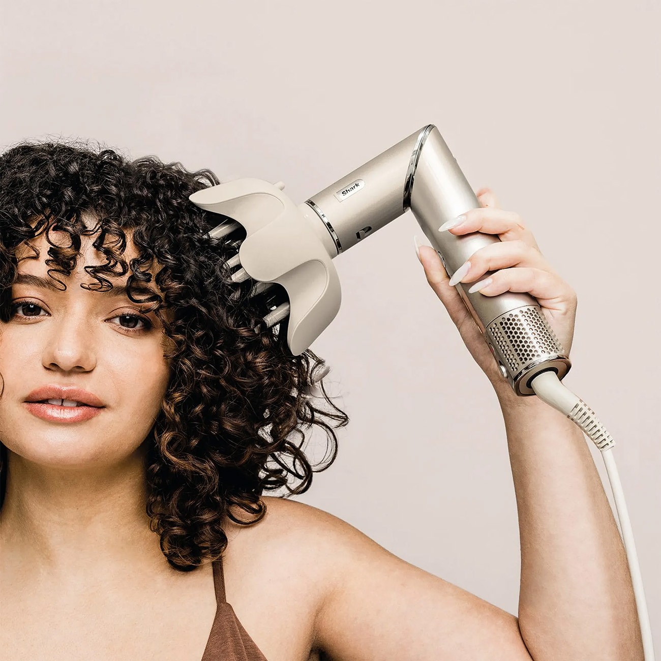 Kobieta używająca suszarki do włosów. Suszarka jest srebrna. Patrzy w kamerę z kręconymi włosami.