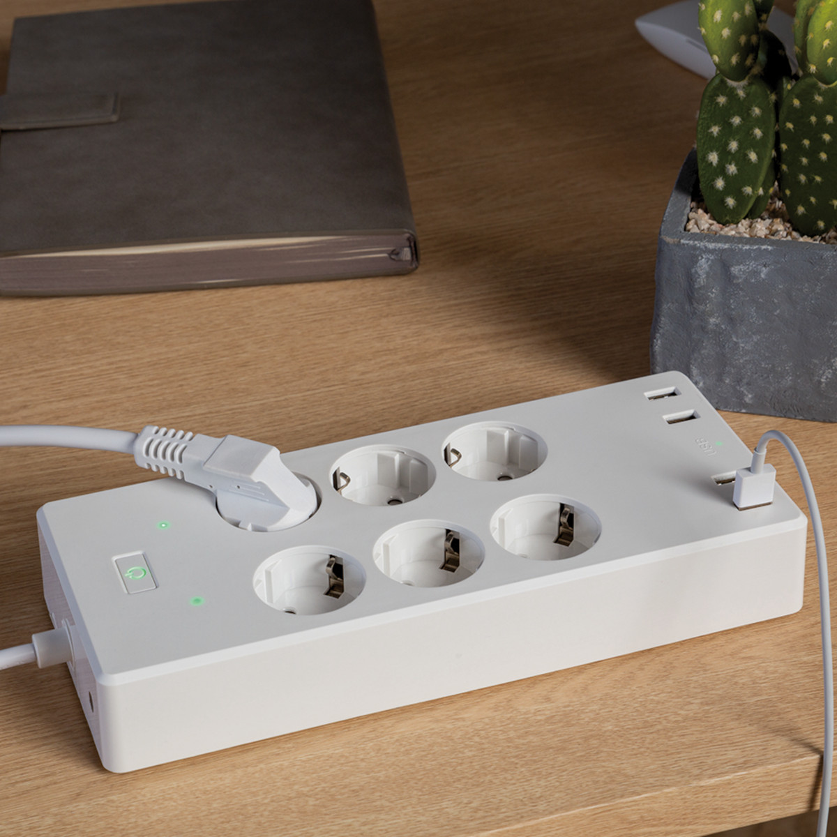 Weiße Steckdosenleiste auf einem Holzschreibtisch mit 5 Steckdosen, 2 USB-Anschlüssen. Notizbuch und Kaktus im Hintergrund.