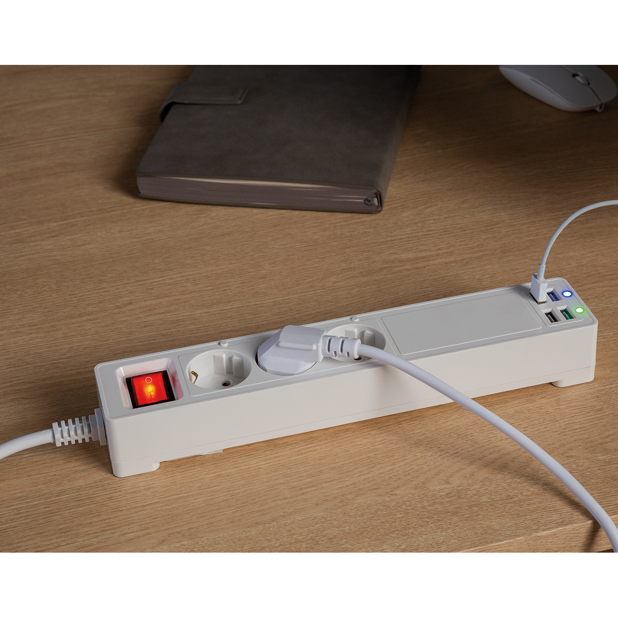 Weiße Steckdosenleiste mit 3 Steckdosen, einem roten Schalter und 4 USB-Anschlüssen. Ein Schreibtisch mit Laptop.