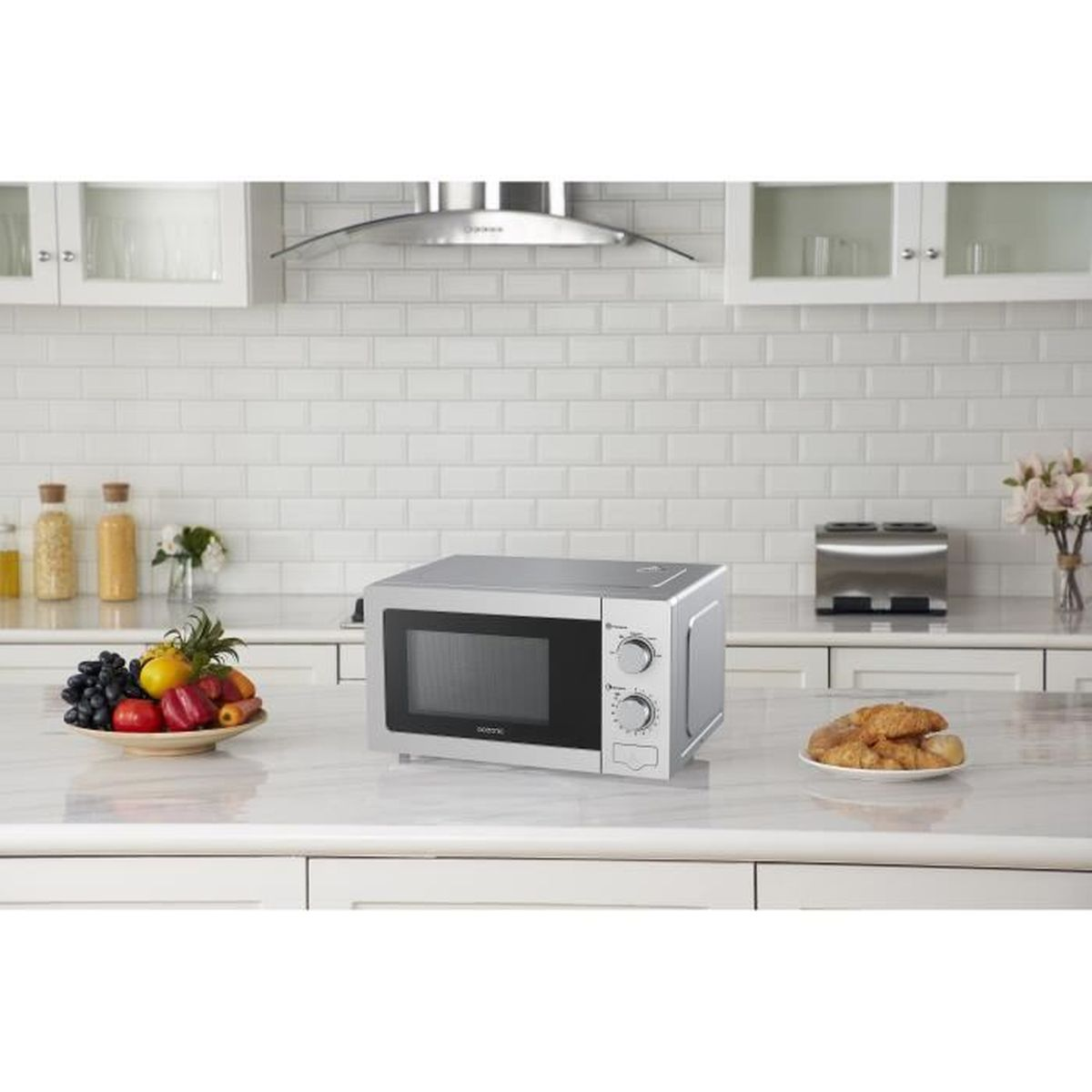 Een zilveren magnetron op een aanrecht met fruit, bloemen en brood.