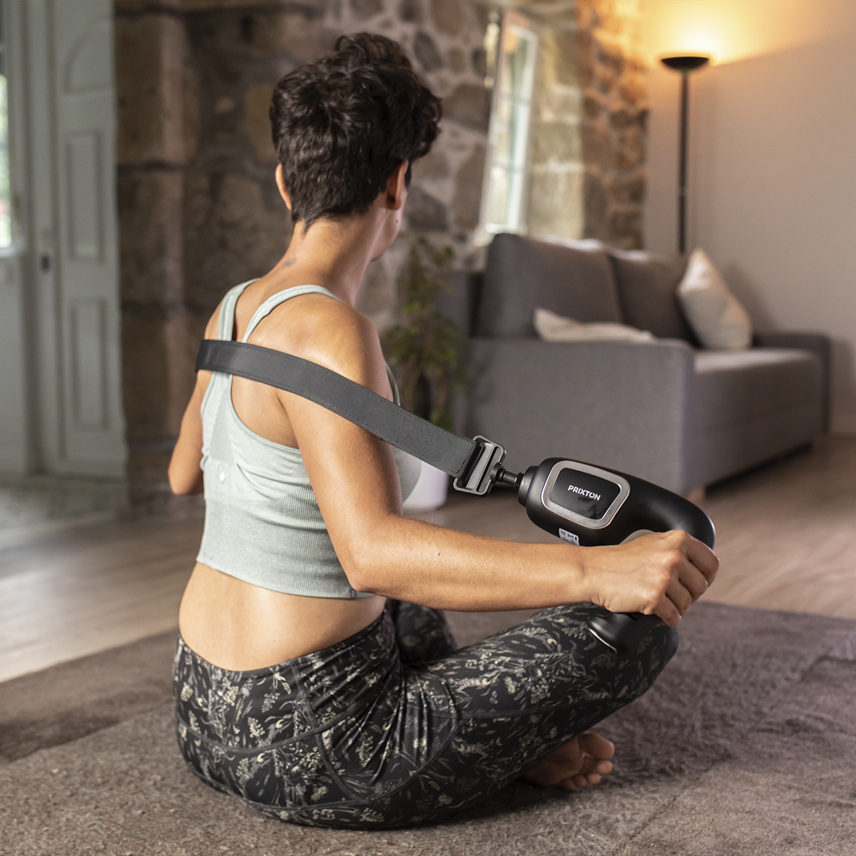 Kobieta używająca masażera pleców siedząc na dywanie. Szara kanapa i lampa w tle.