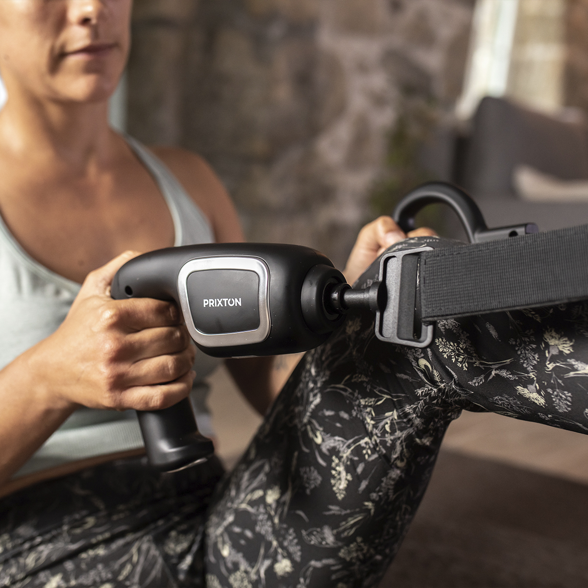 Osoba używająca czarnego pistoletu do masażu z paskiem. Na pierwszym planie urządzenie i dłonie, w tle pokój.