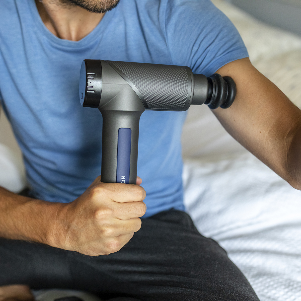 Osoba używa pistoletu do masażu na ramieniu. Widoczna niebieska koszula i białe łóżko.