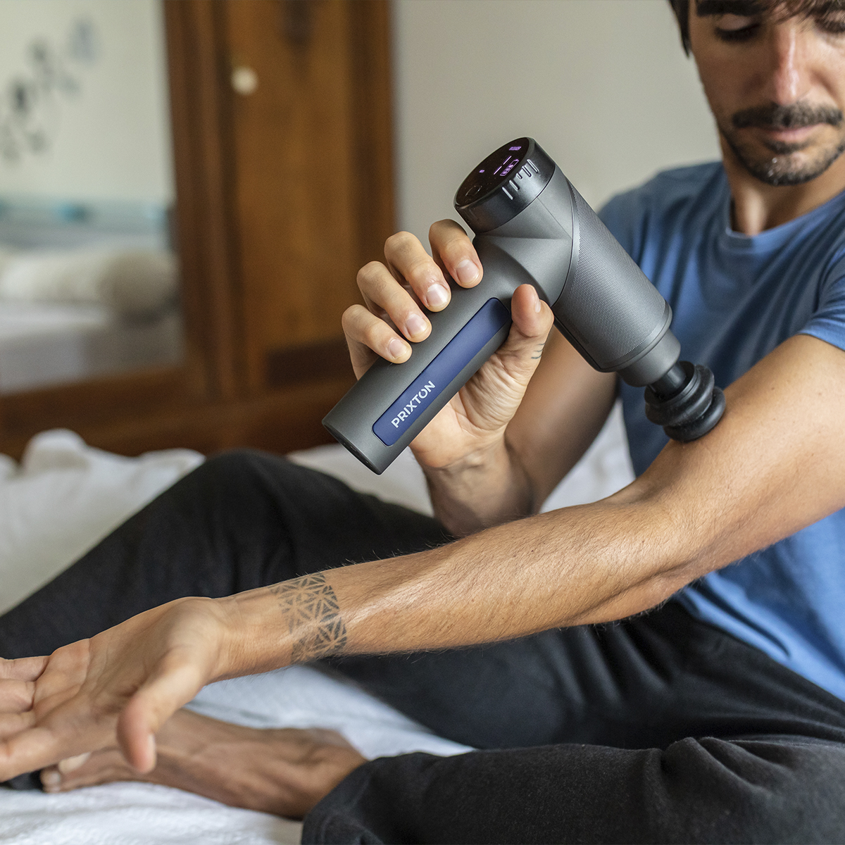 Mężczyzna używa pistoletu do masażu na ramieniu. Widoczny tatuaż. W tle łóżko.