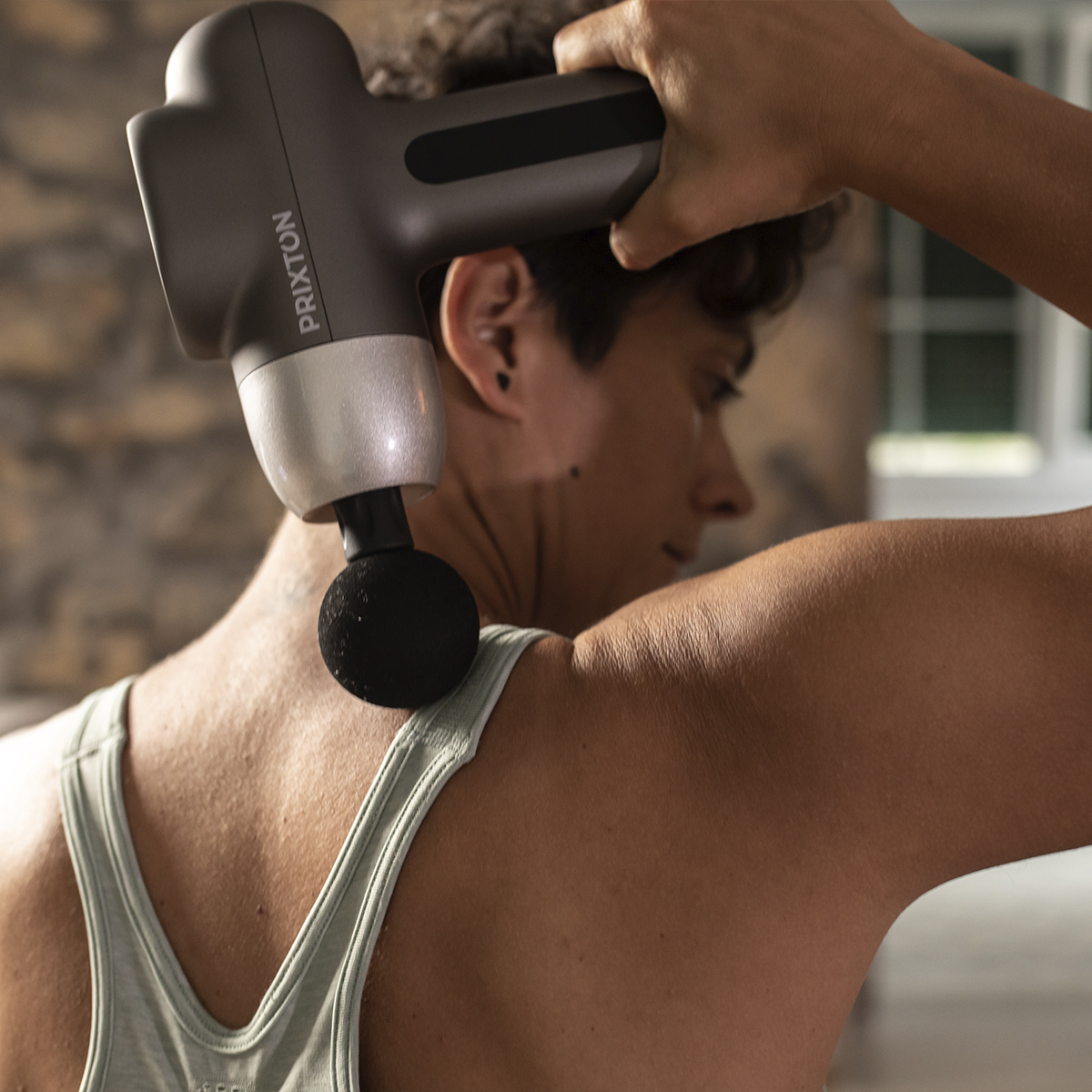 Osoba używa pistoletu do masażu na plecach. Pistolet jest szary, osoba ma na sobie zielony podkoszulek.