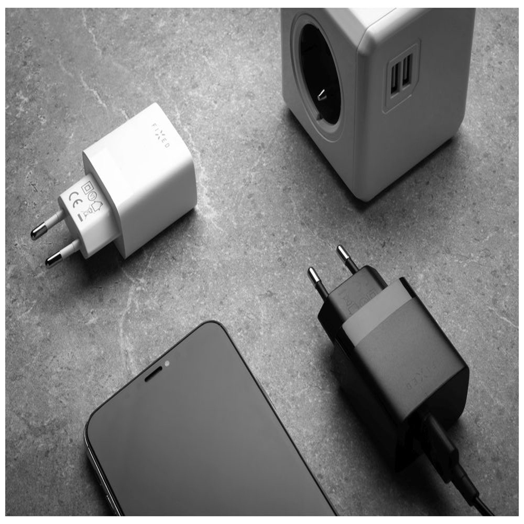 Schwarz-Weiß-Bild eines Smartphones, Ladegeräts und Steckdosenadapters auf strukturierter Oberfläche.