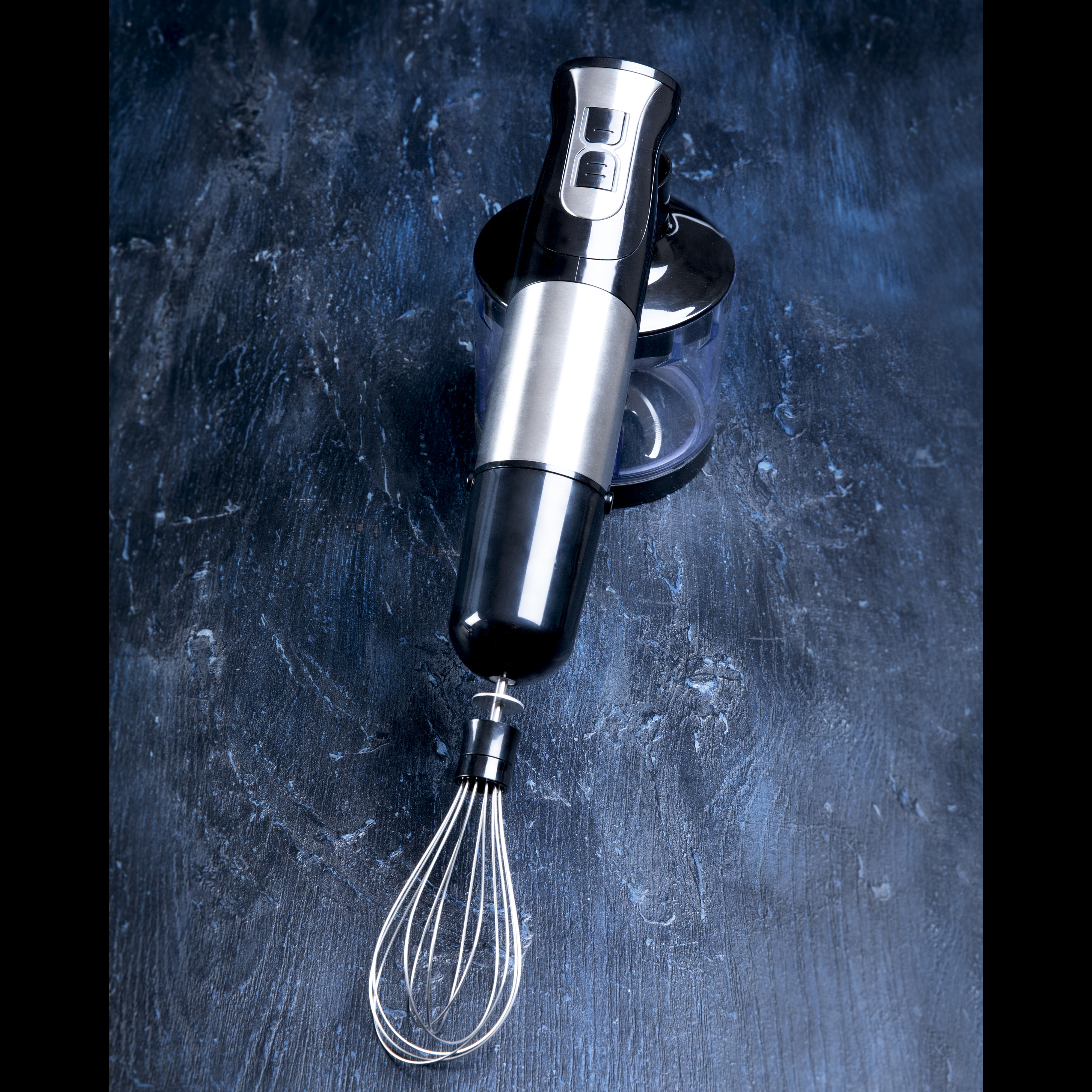 Czarny i srebrny blender ręczny z końcówką do ubijania na ciemnoniebieskiej powierzchni.