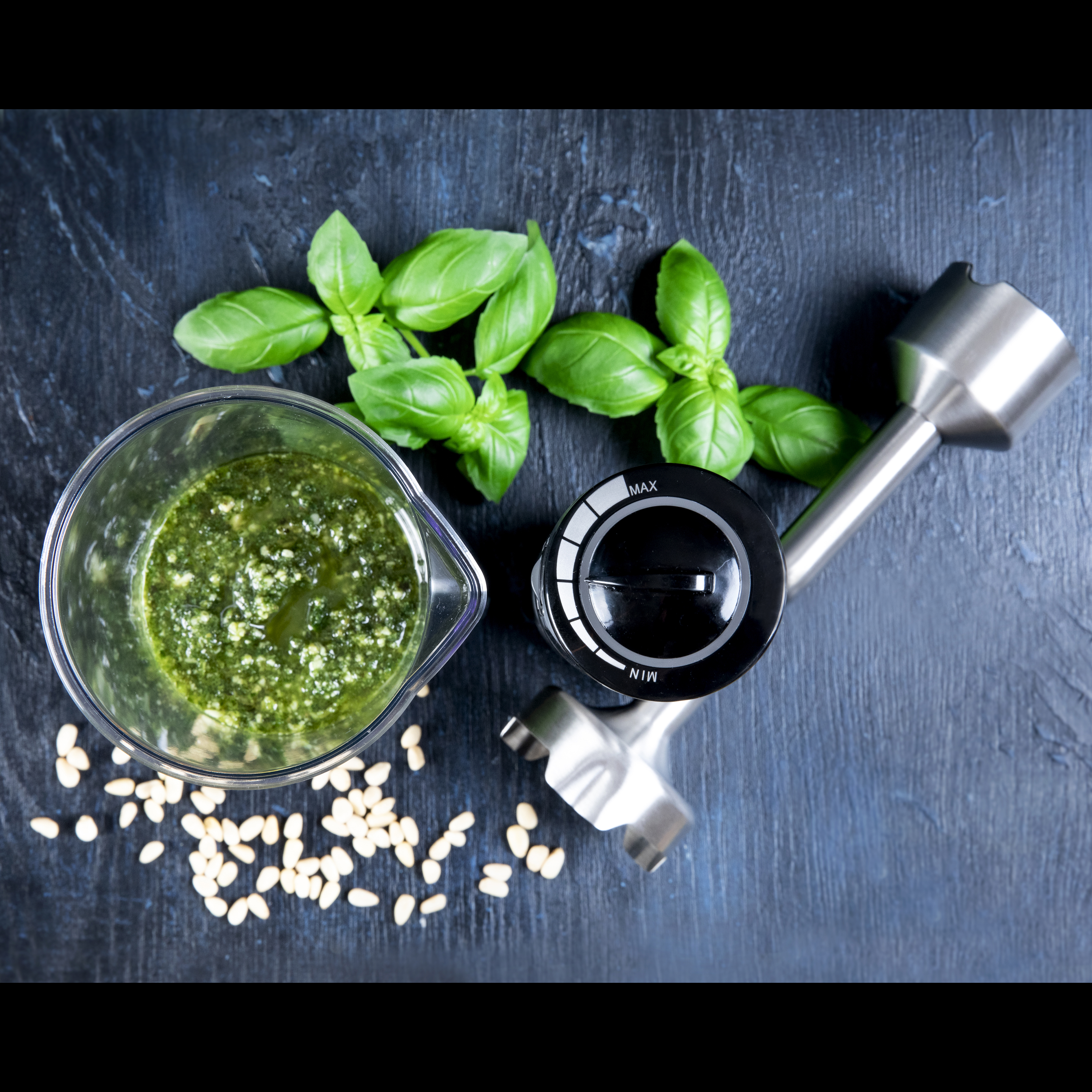 Widok z góry na pesto w szklance, liście bazylii i orzeszki piniowe na ciemnoniebieskiej powierzchni.