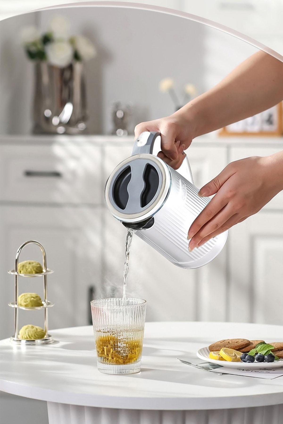Eine Person gießt Wasser aus einem weißen Wasserkocher in ein Glas. Kekse und Obst sind auf dem Tisch.