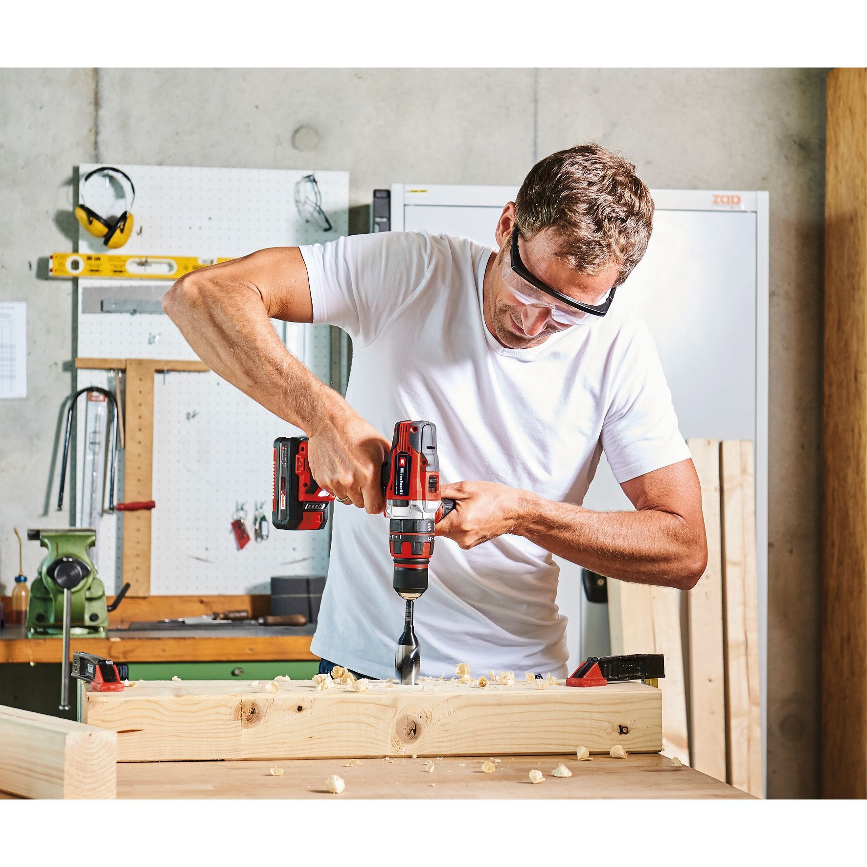 Mężczyzna w okularach ochronnych wierci w drewnie czerwoną wiertarką akumulatorową.