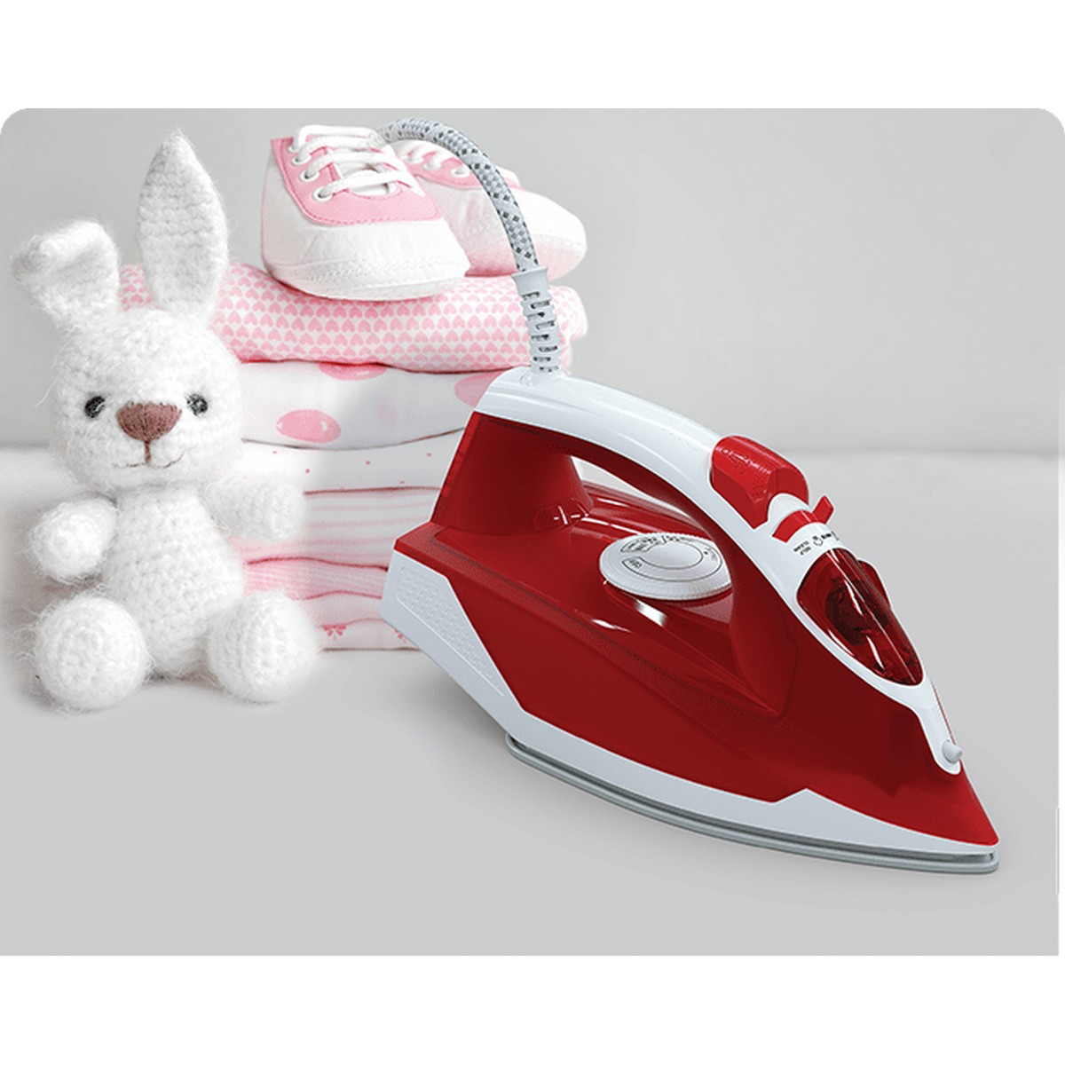 Plancha roja sobre superficie blanca. Conejo de juguete, zapatos y ropa de bebé cerca.