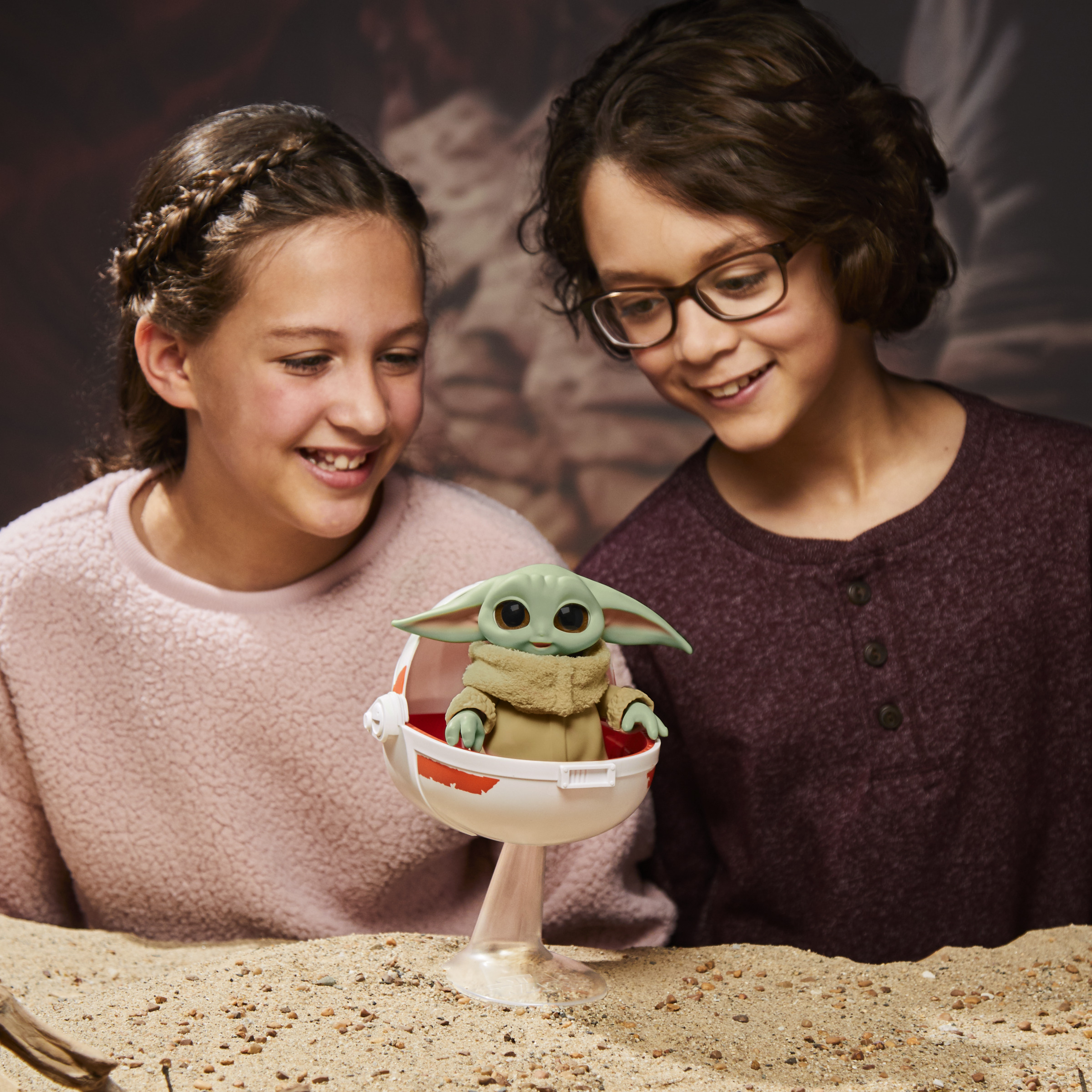 Dos niños mirando un juguete de Baby Yoda en una cápsula. Están sonriendo. Arena marrón y un fondo.
