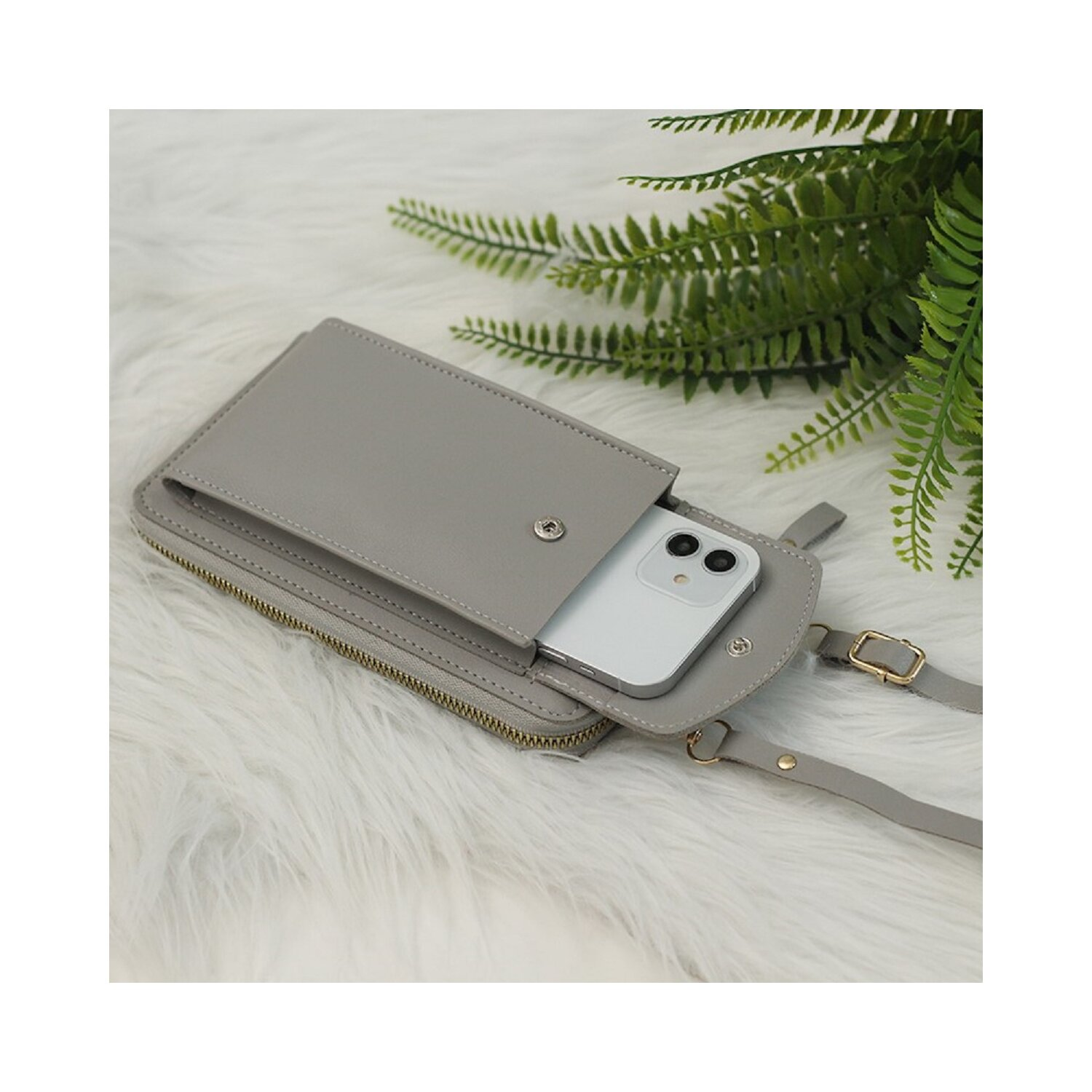 Eine graue Umhängetasche mit einem Telefon darin, platziert auf einer flauschigen weißen Oberfläche mit grünen Blättern.
