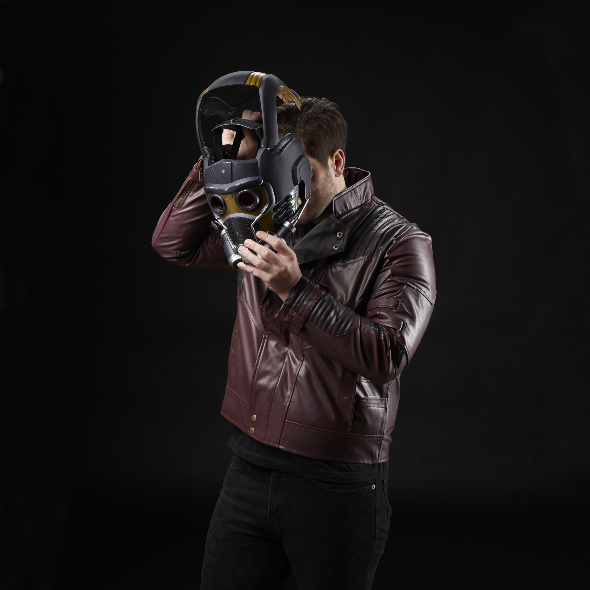 Hombre con chaqueta de cuero roja sosteniendo un casco de Star-Lord contra fondo negro.