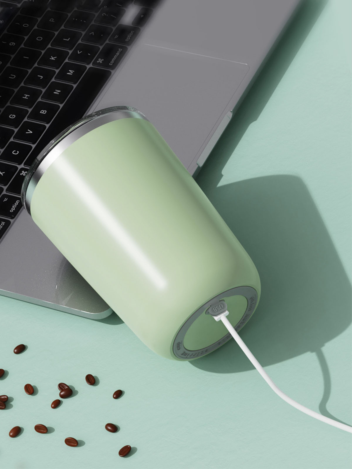 Ein grüner Becher steht auf einem Laptop, verbunden mit einem weißen Ladekabel, mit Kaffeebohnen drumherum.