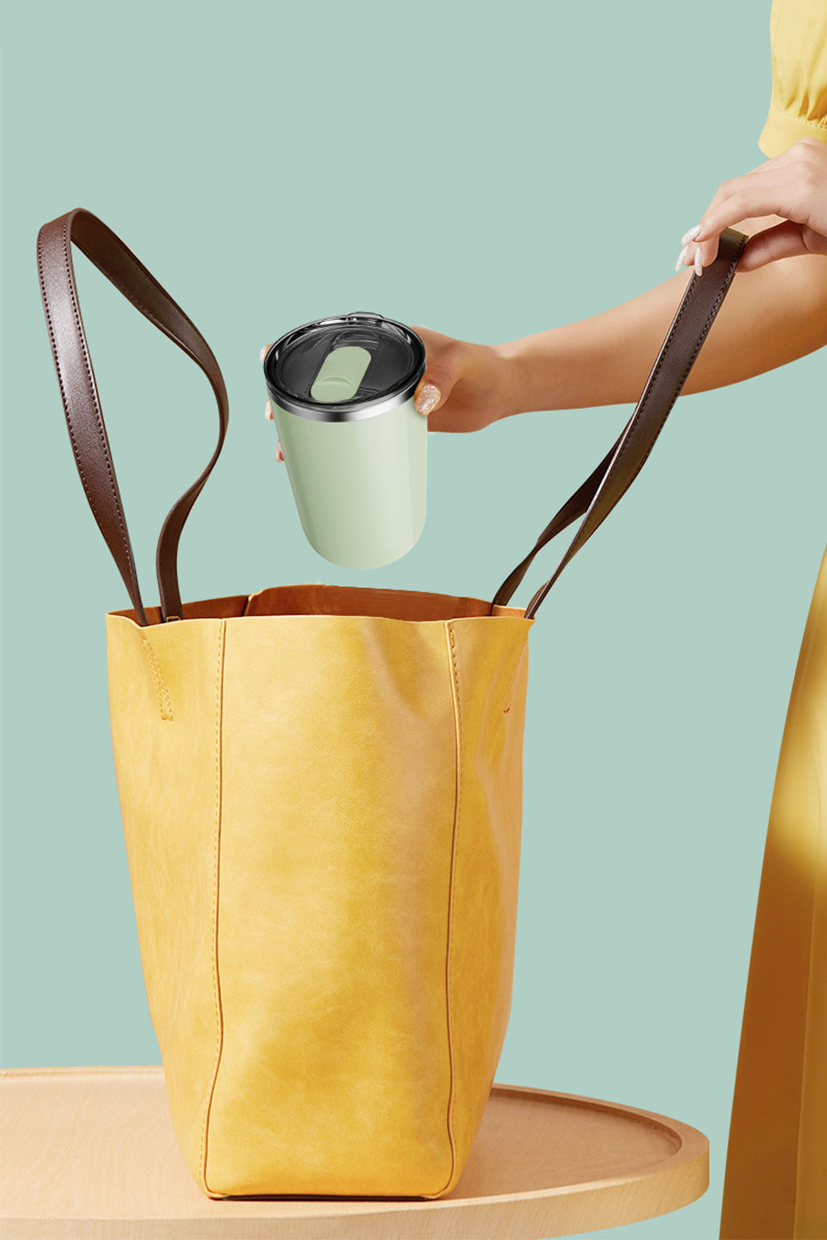 Eine Person legt einen hellgrünen Becher mit schwarzem Deckel in eine gelbe Handtasche.