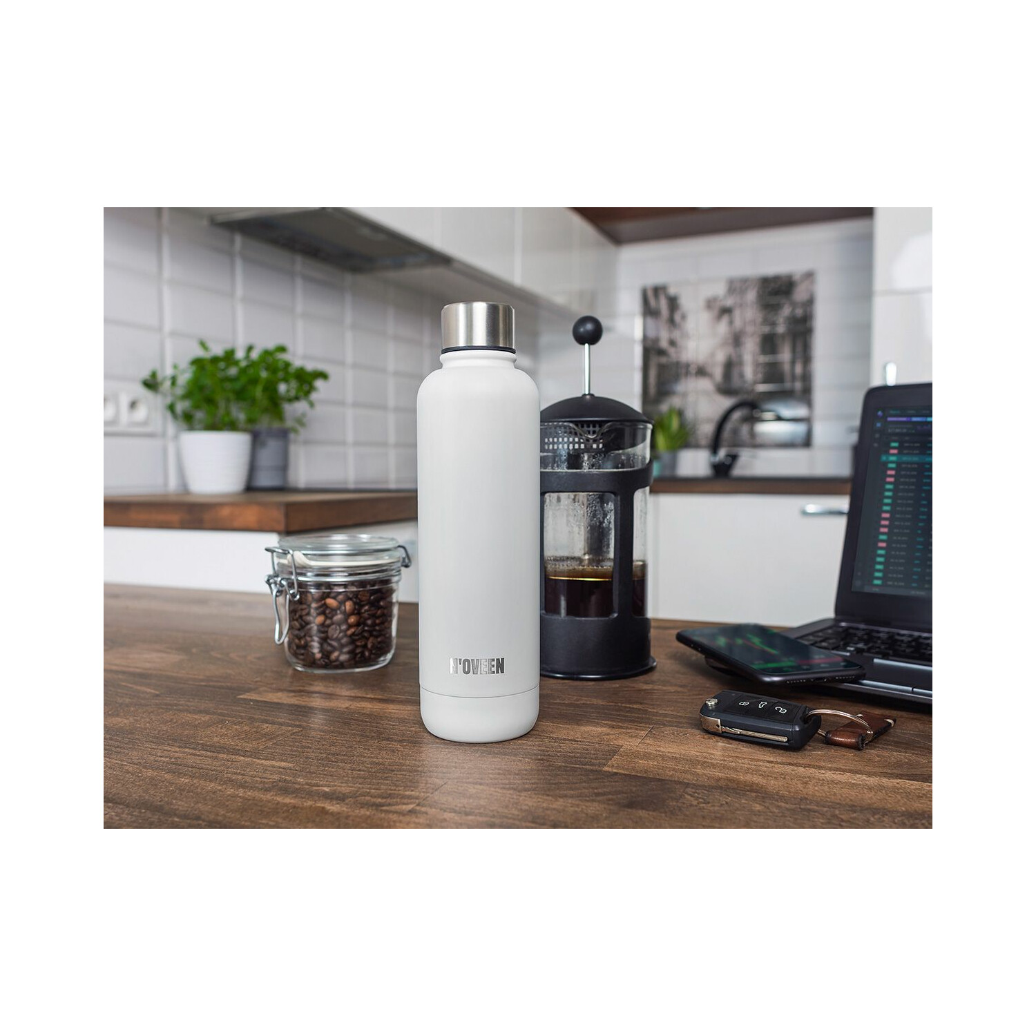 Weiße Flasche, Kaffeepresse und Laptop auf einem Holztisch. Hintergrund zeigt Küche mit Pflanzen.