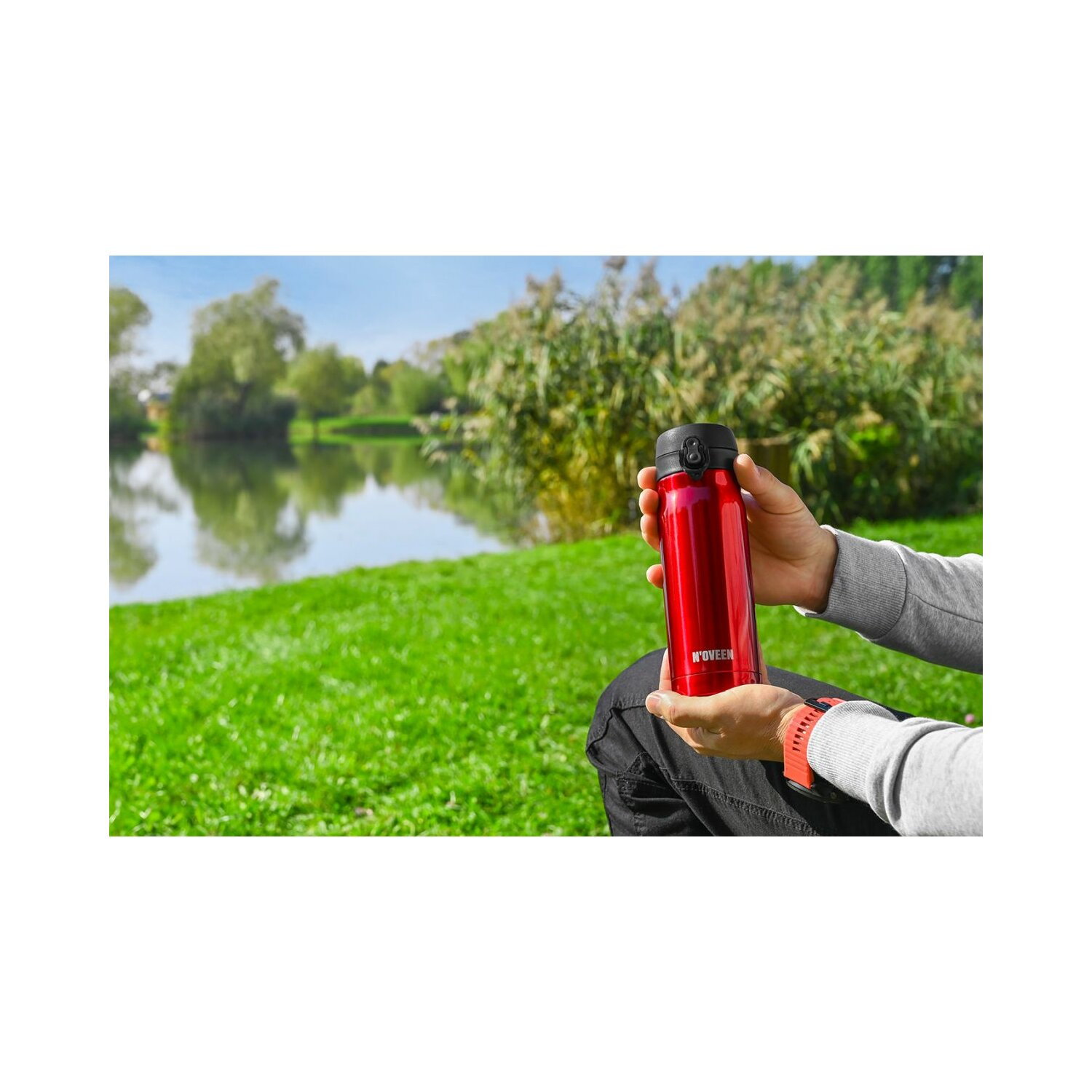 Person sitzt am See, hält rote Wasserflasche. Grünes Gras im Vordergrund, Bäume im Hintergrund.