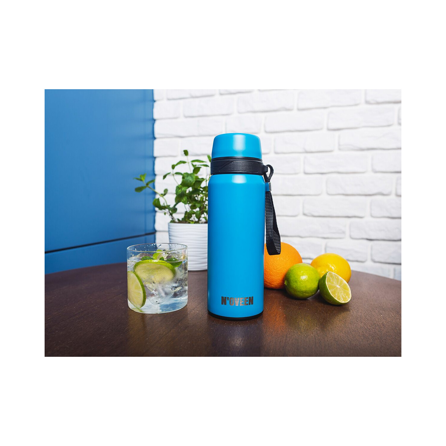 Eine blaue Wasserflasche, ein Glas mit Limettenscheiben und Früchte auf einem Tisch mit weißer Backsteinmauer.