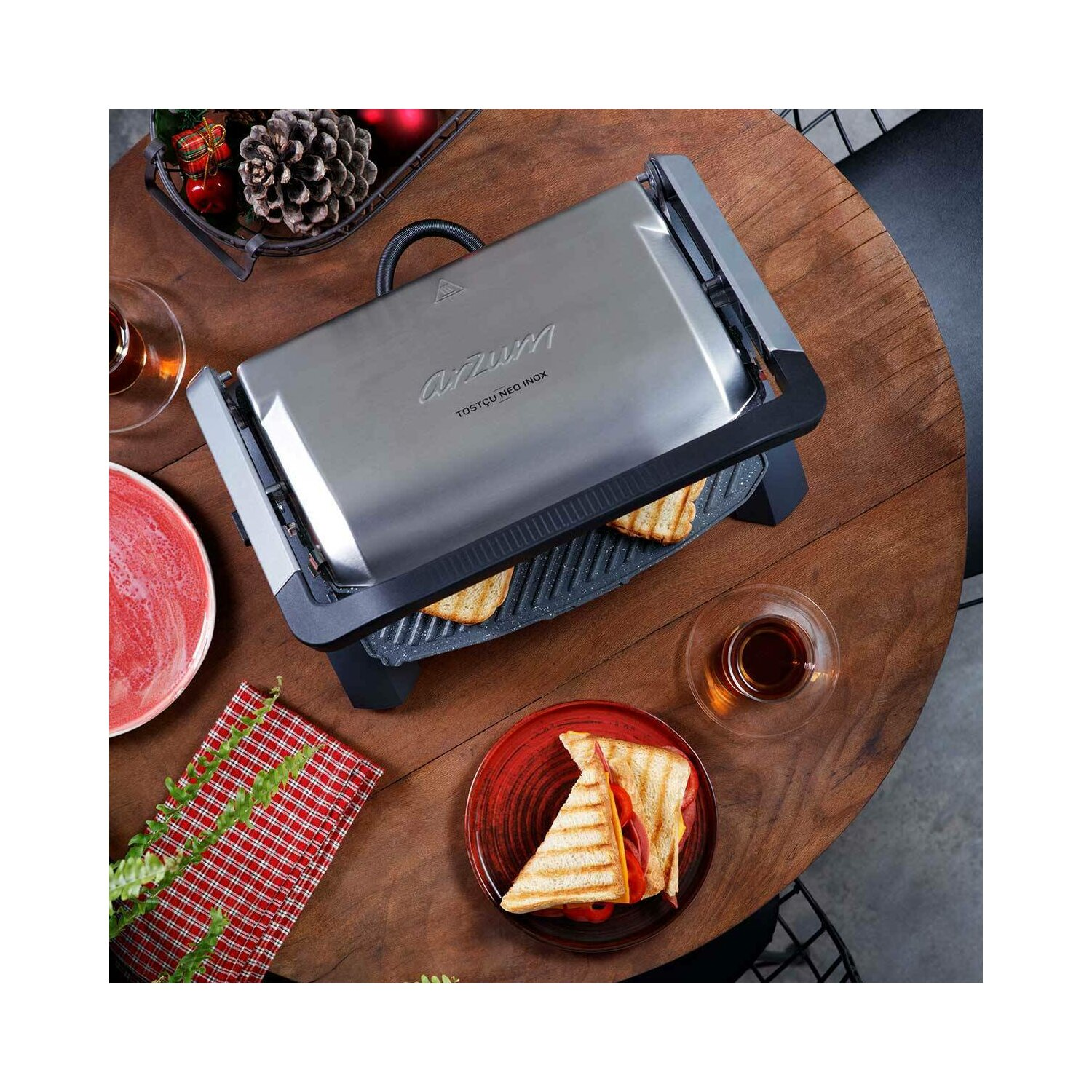 Ein Sandwich-Toaster mit Sandwiches auf einem Holztisch, mit einem roten Teller und einer Tasse Tee.