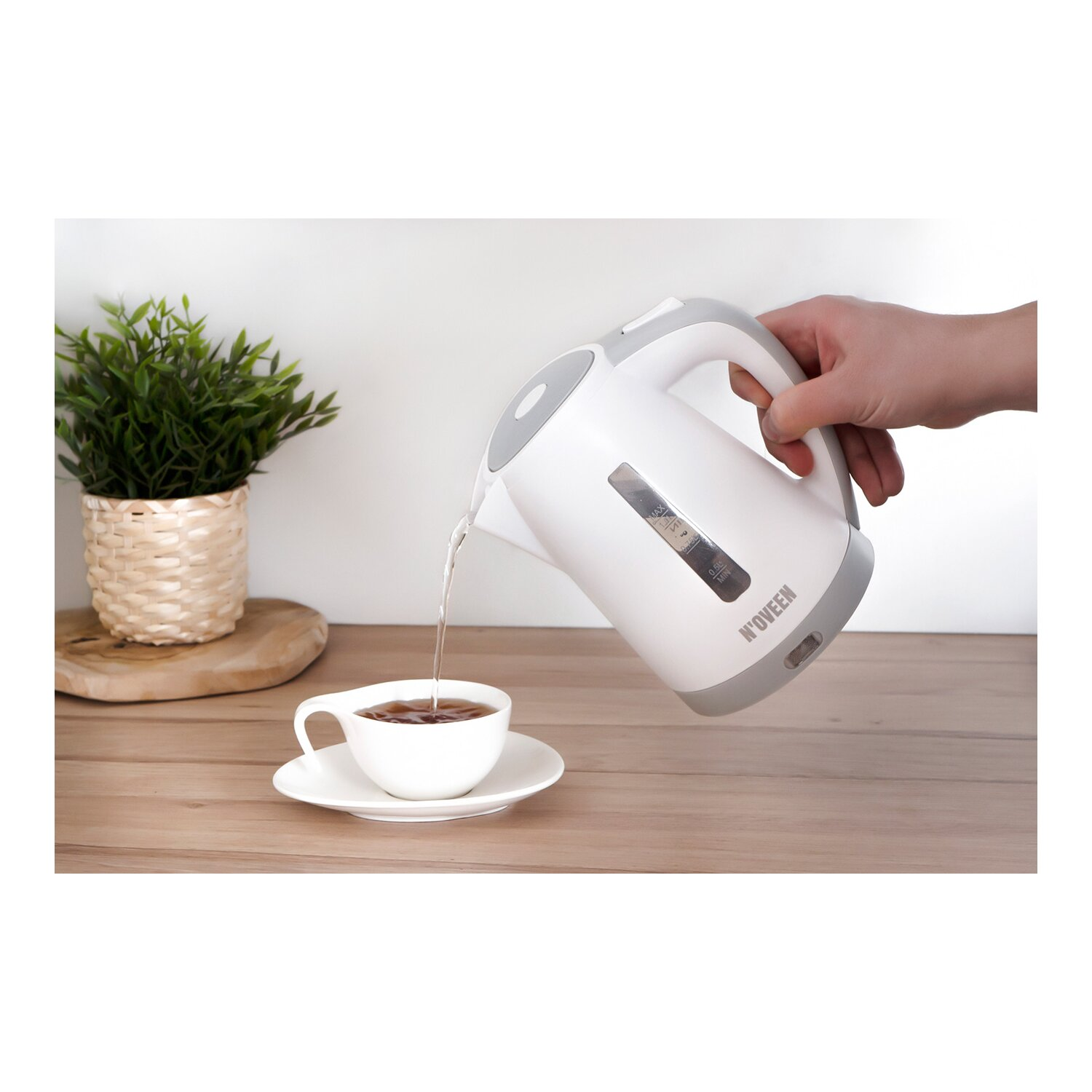 Biały czajnik elektryczny wlewa wodę do białej filiżanki na spodku. Roślina jest w tle.