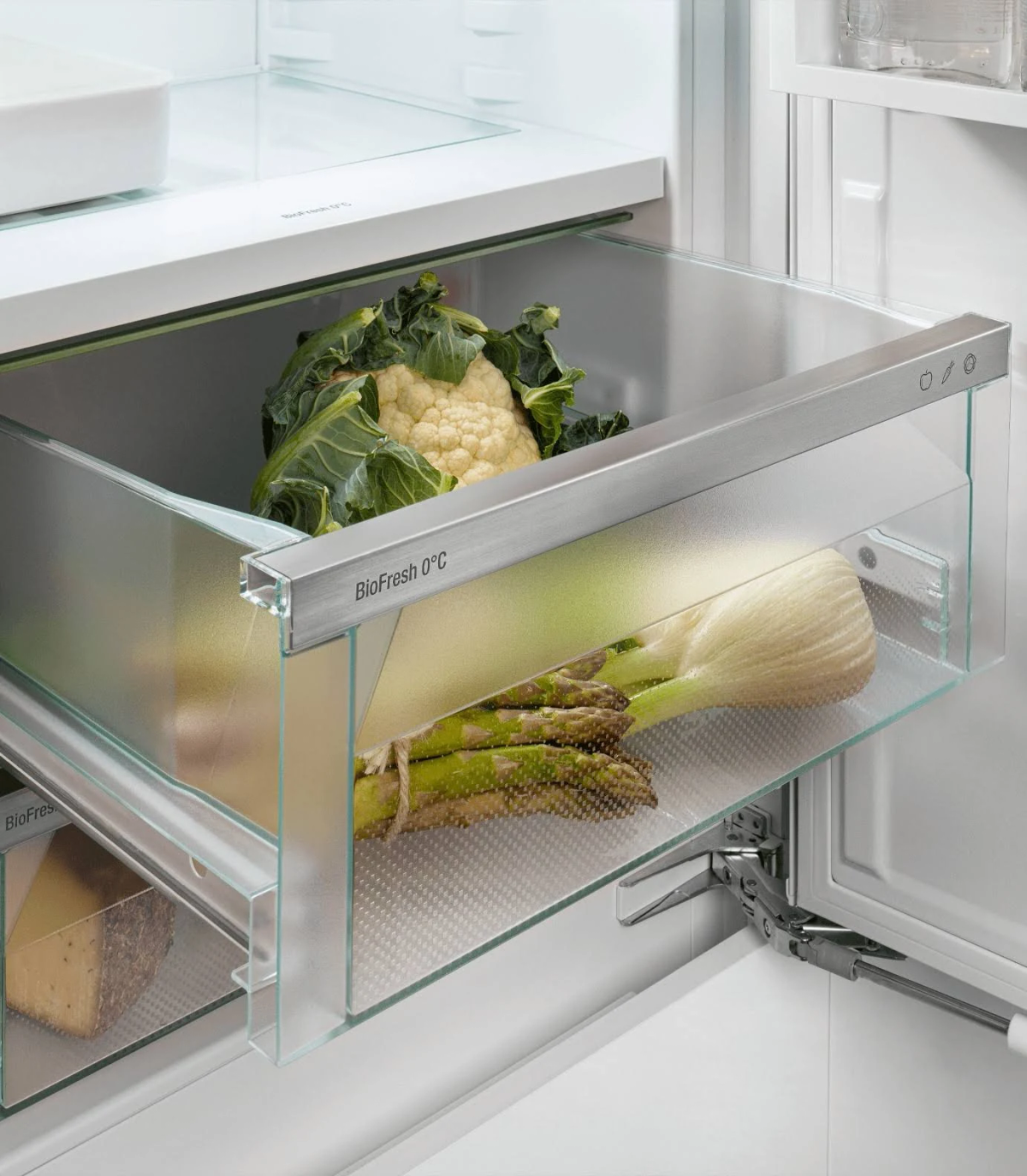 Un cajón transparente dentro de un refrigerador, lleno de verduras. Incluye espárragos y coliflor.