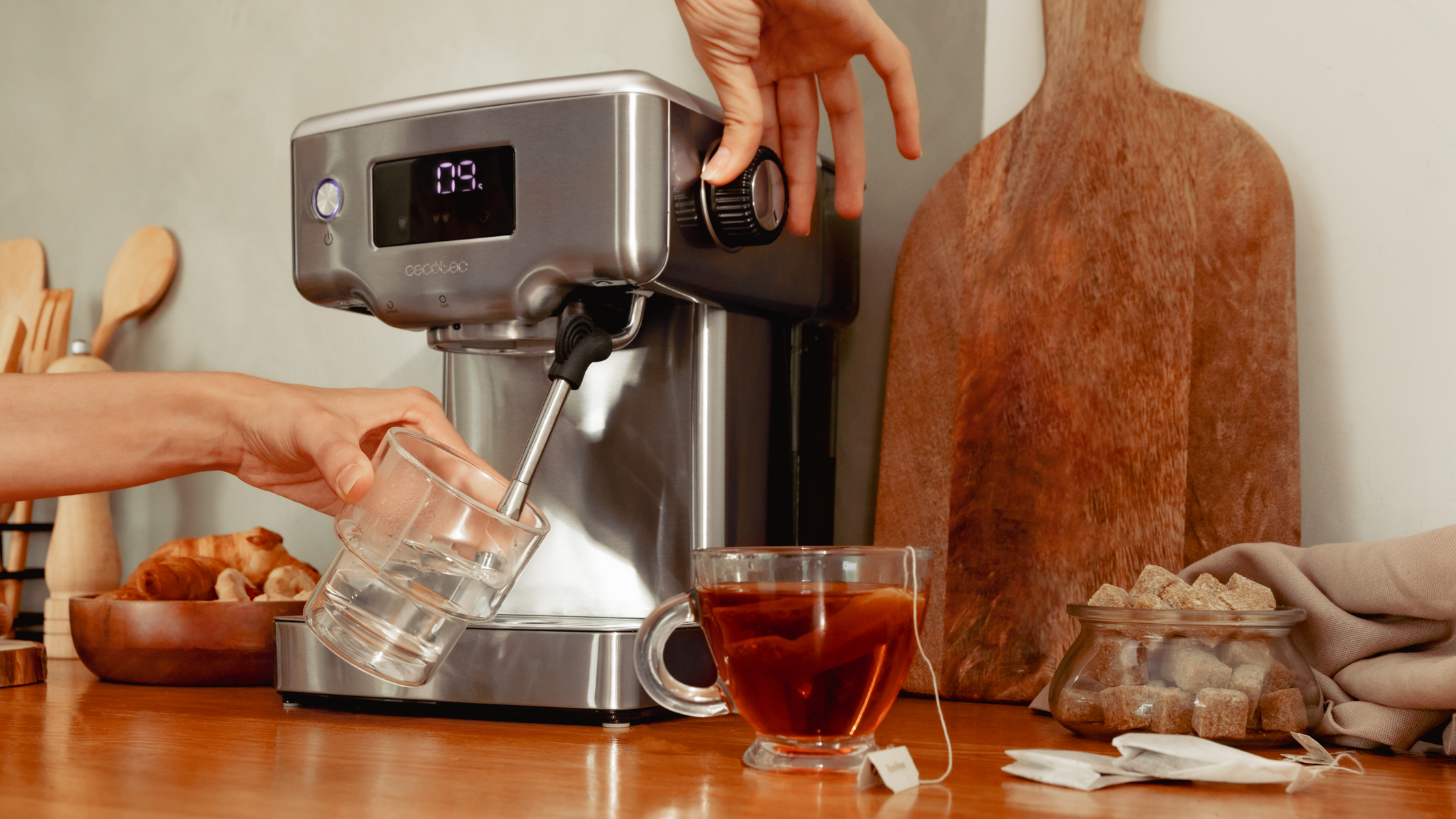 Osoba nalewa wodę do szklanki ze srebrnego ekspresu do kawy. Widoczna jest filiżanka herbaty.