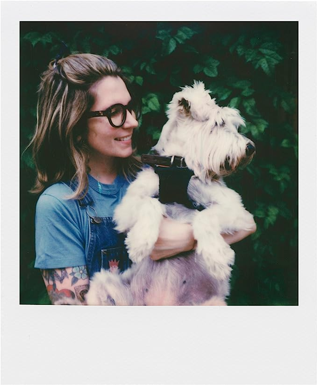 Kobieta w okularach i kombinezonie uśmiecha się, trzymając puszystego białego psa przed zielonym krzakiem.
