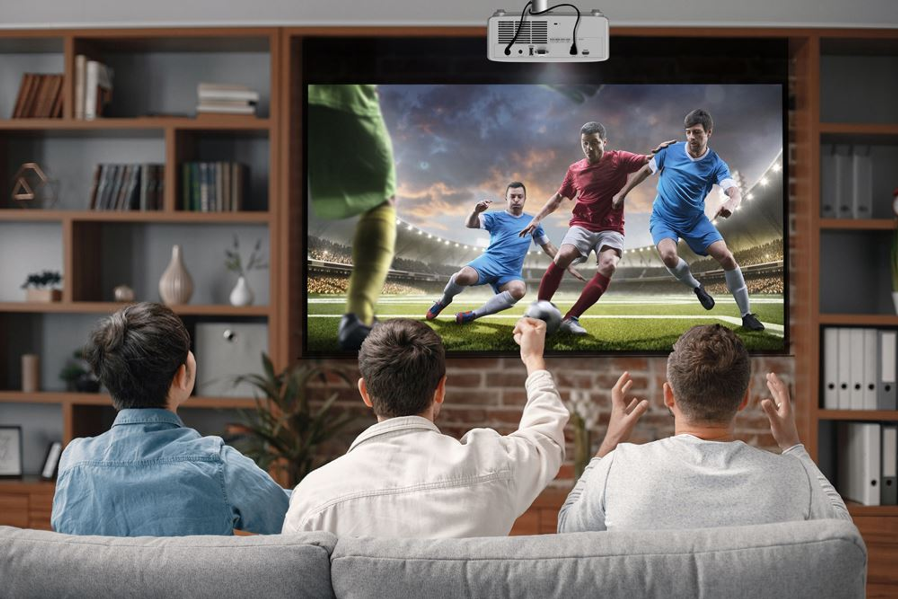 Trzy osoby oglądające mecz piłki nożnej na dużym ekranie, kibicujące i podekscytowane.