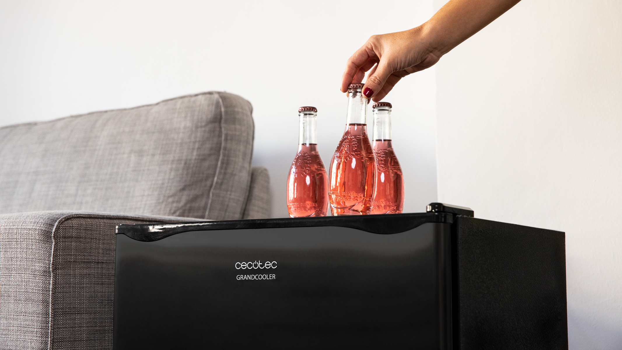 Una mano che posiziona una bottiglia sopra un mini-frigo nero. Sono visibili tre bottiglie color rosa.