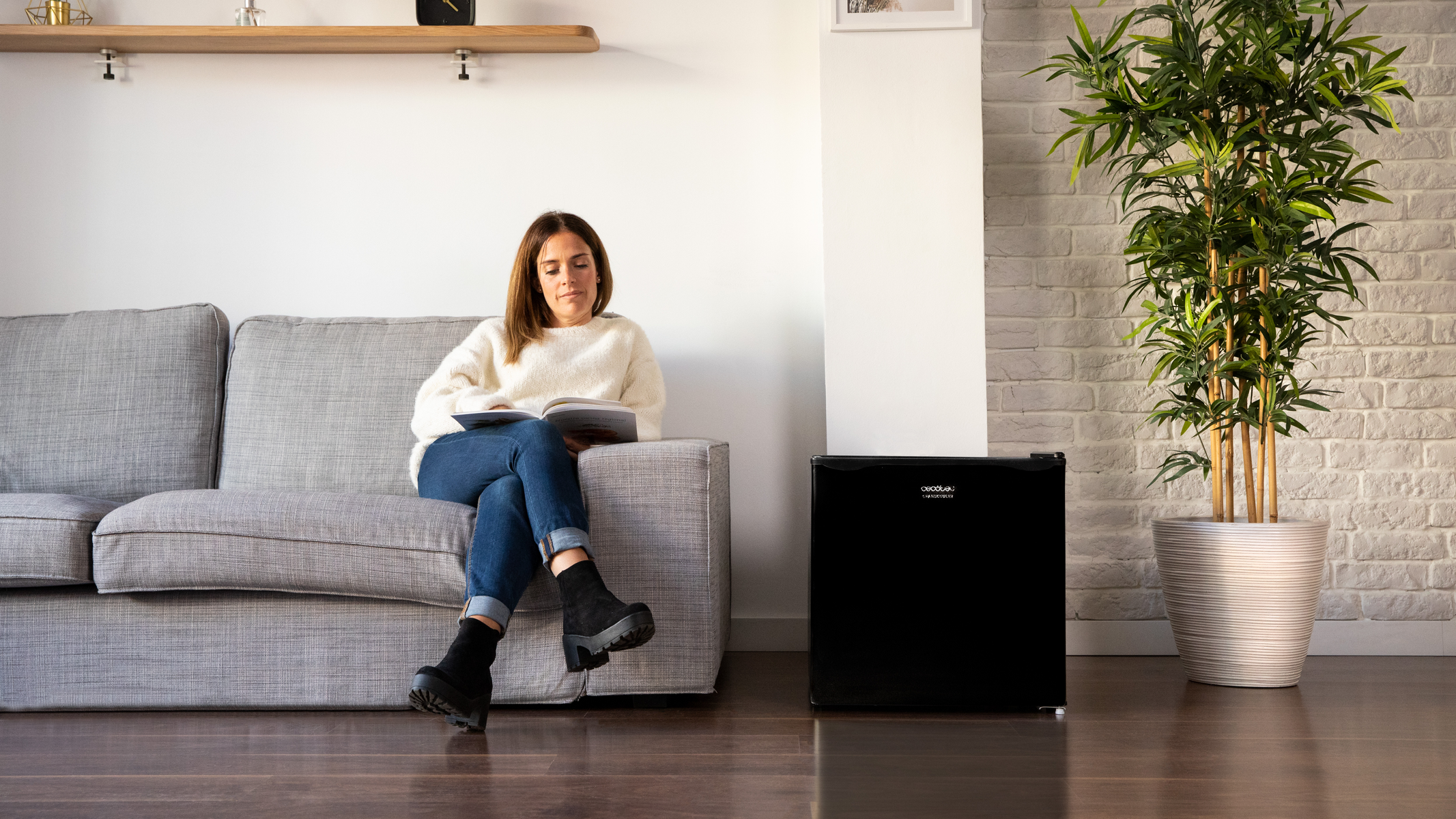 Una donna legge un libro su un divano grigio, con un mini frigo nero e una pianta nelle vicinanze.