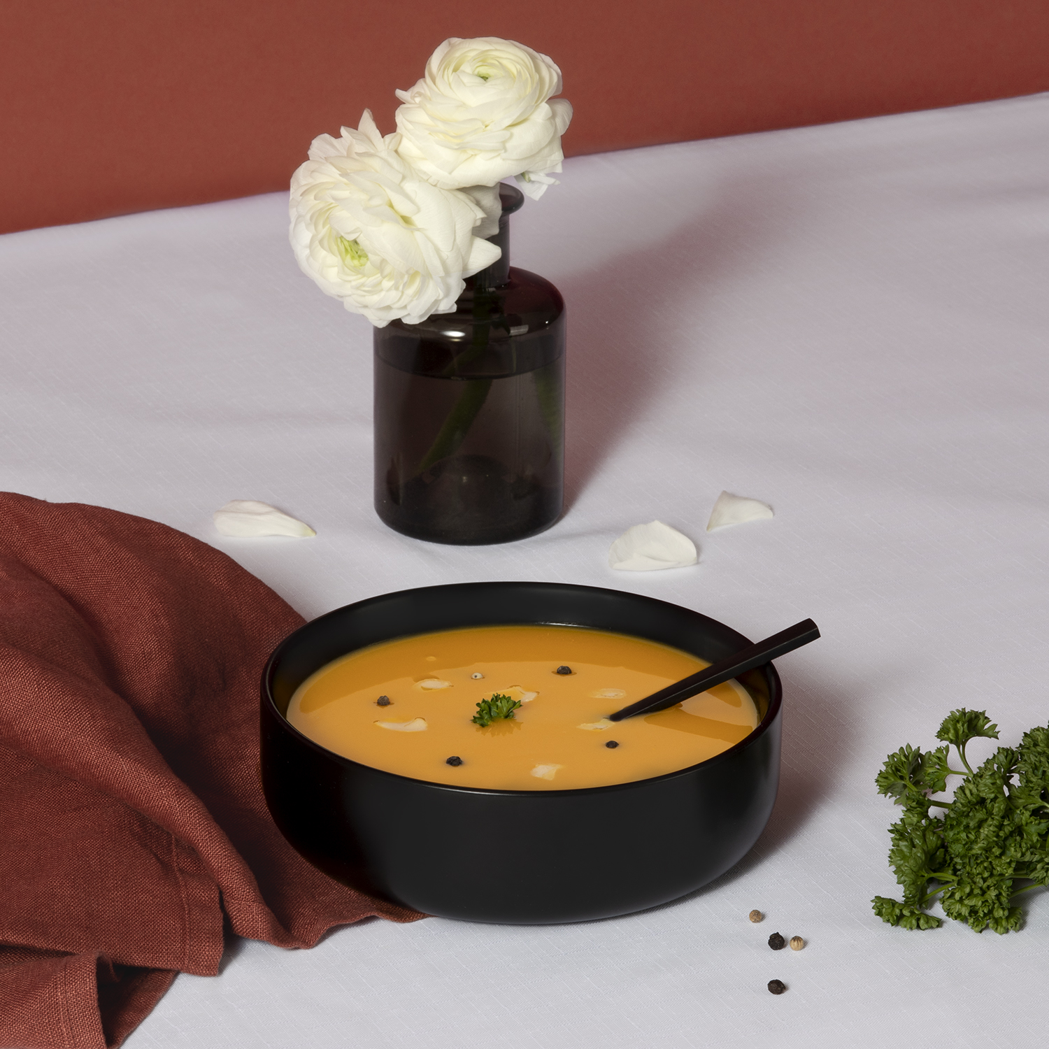 Zuppa arancione in ciotola nera, fiori bianchi in vaso, stoffa rossa su tavolo bianco.
