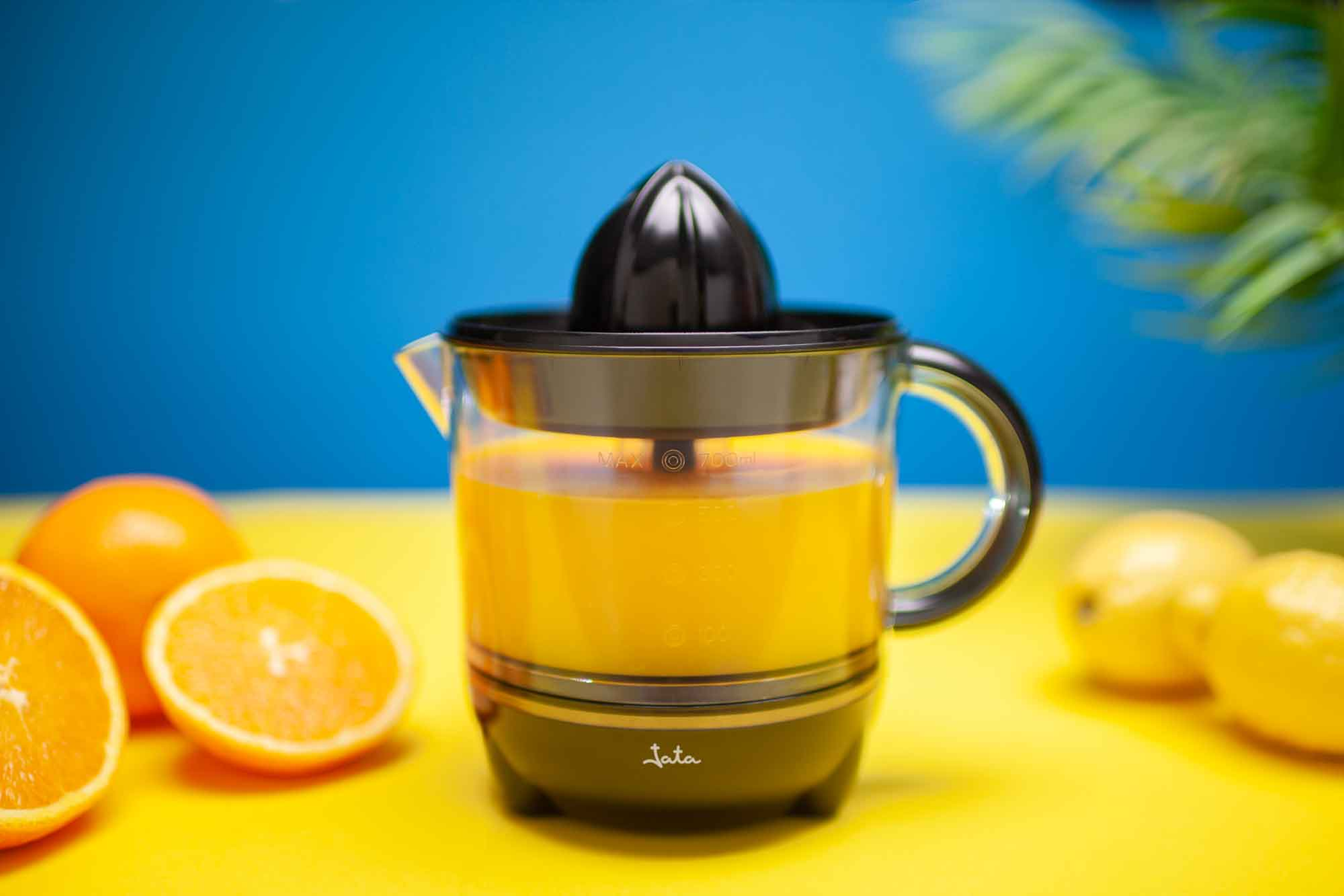 Una máquina de zumo con zumo de naranja, naranjas y limones sobre una superficie amarilla contra un fondo azul.