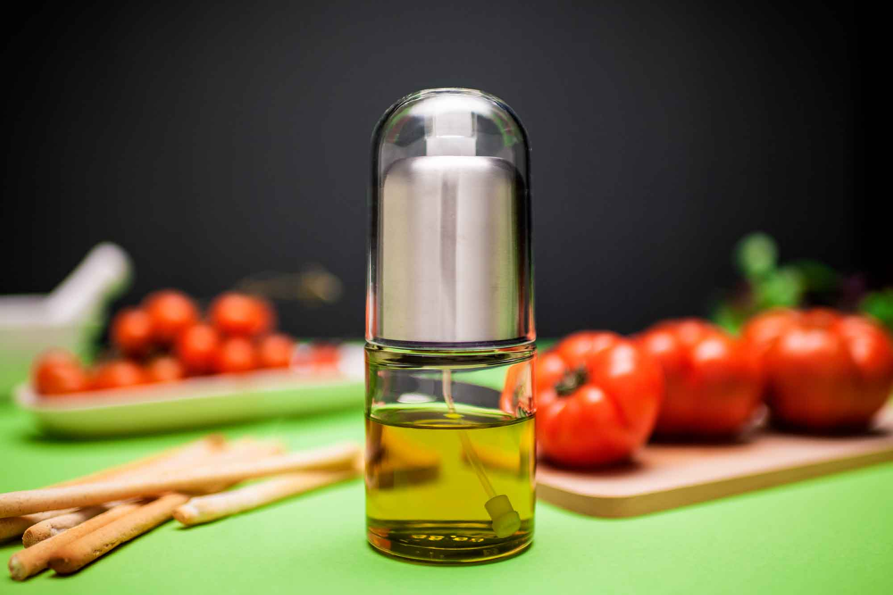 Pulverizador de aceite con aceite de oliva, tomates y palitos de pan sobre mesa verde y fondo negro.