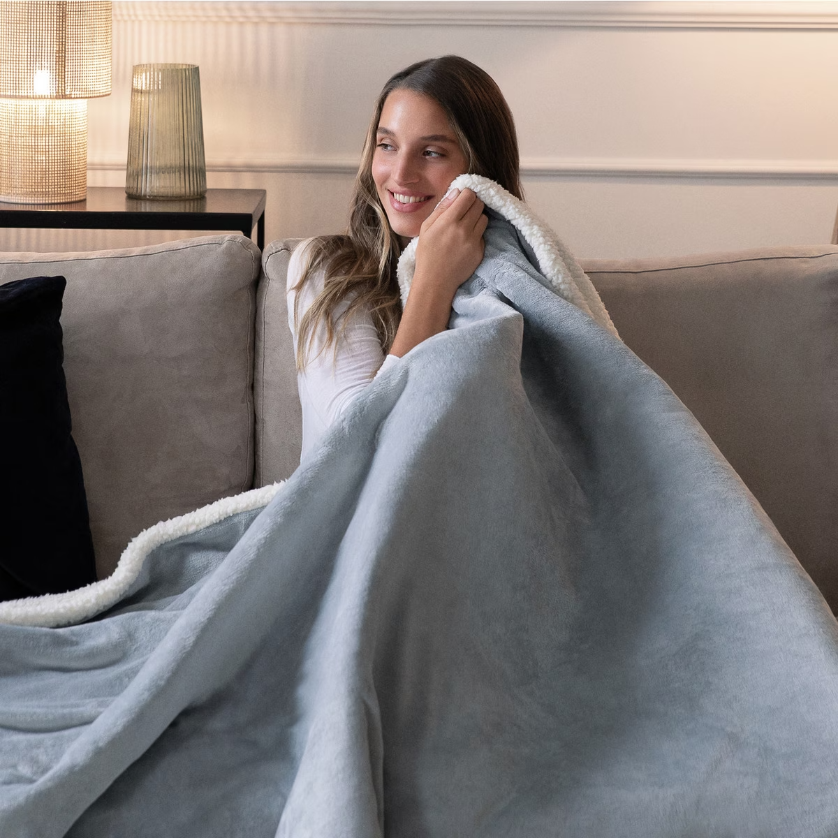 Una mujer sonriente sentada en un sofá, envuelta en una manta azul claro con ribete blanco.