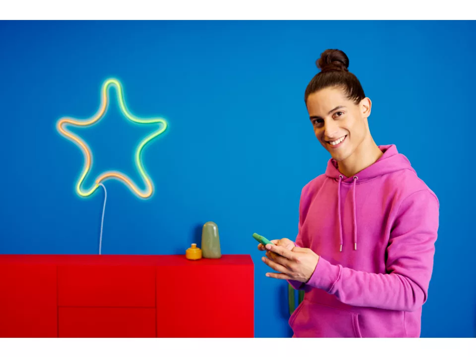 Uomo che tiene un telefono sorridendo, davanti a mobile rosso e muro blu.