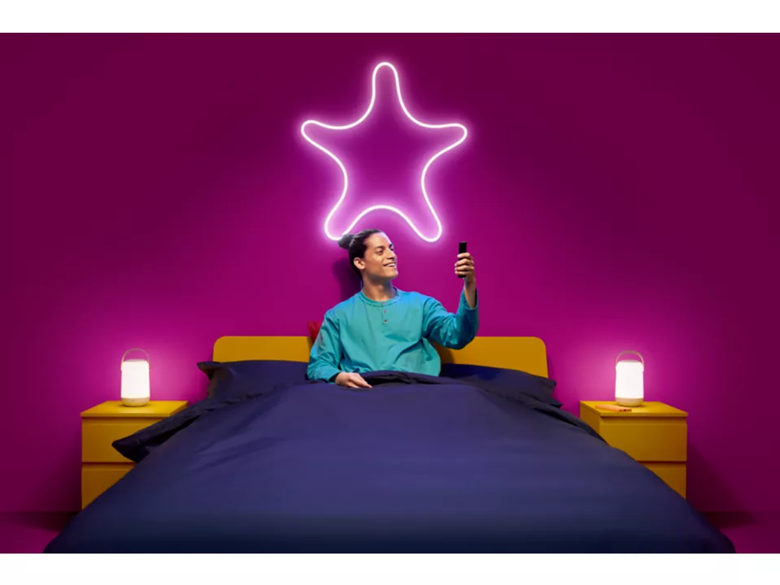 Uomo a letto che guarda il telefono, stella al neon sopra. Due lampade sui comodini. Muro viola, letto giallo.