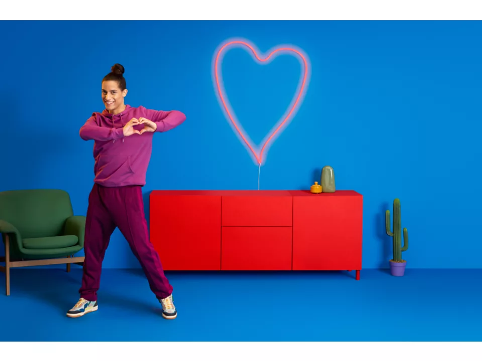 Donna fa una forma a cuore. Un mobile rosso e un cuore al neon dietro di lei.