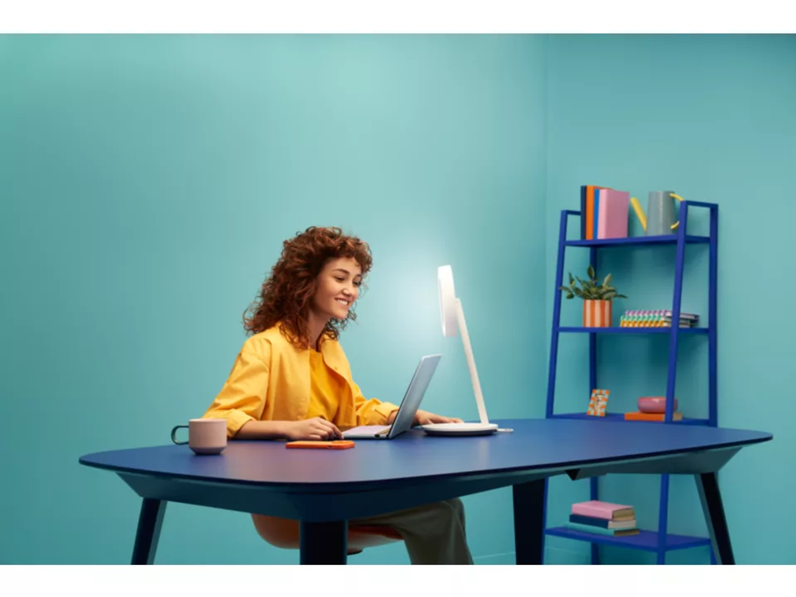 Kobieta używająca laptopa przy niebieskim biurku. Nad nią światło, a w tle półka i roślina.