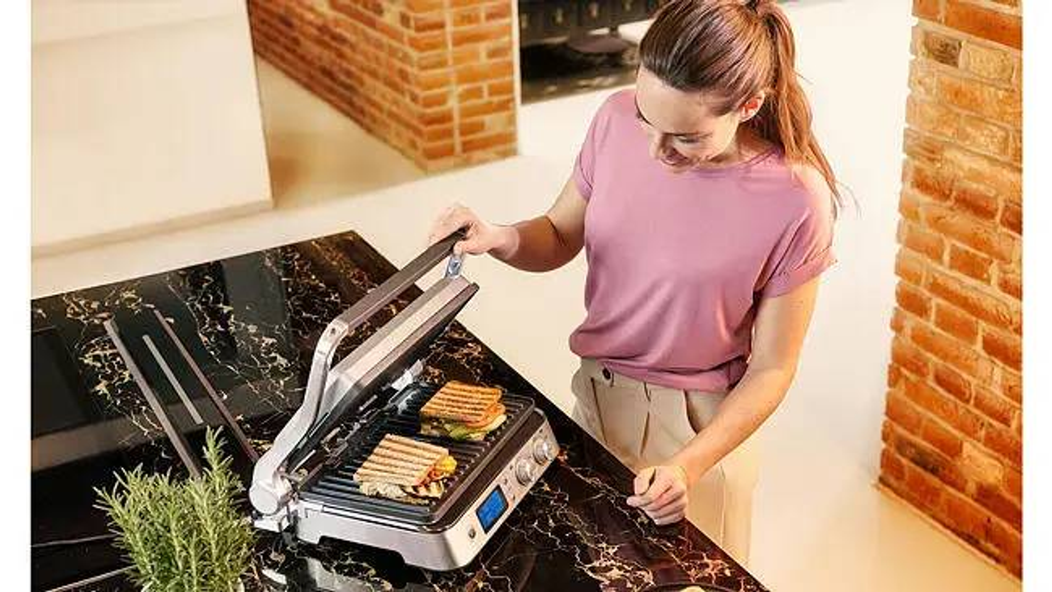 Frau benutzt eine Panini-Presse auf einer schwarzen Marmor-Arbeitsplatte. Zwei Sandwiches sind drin.