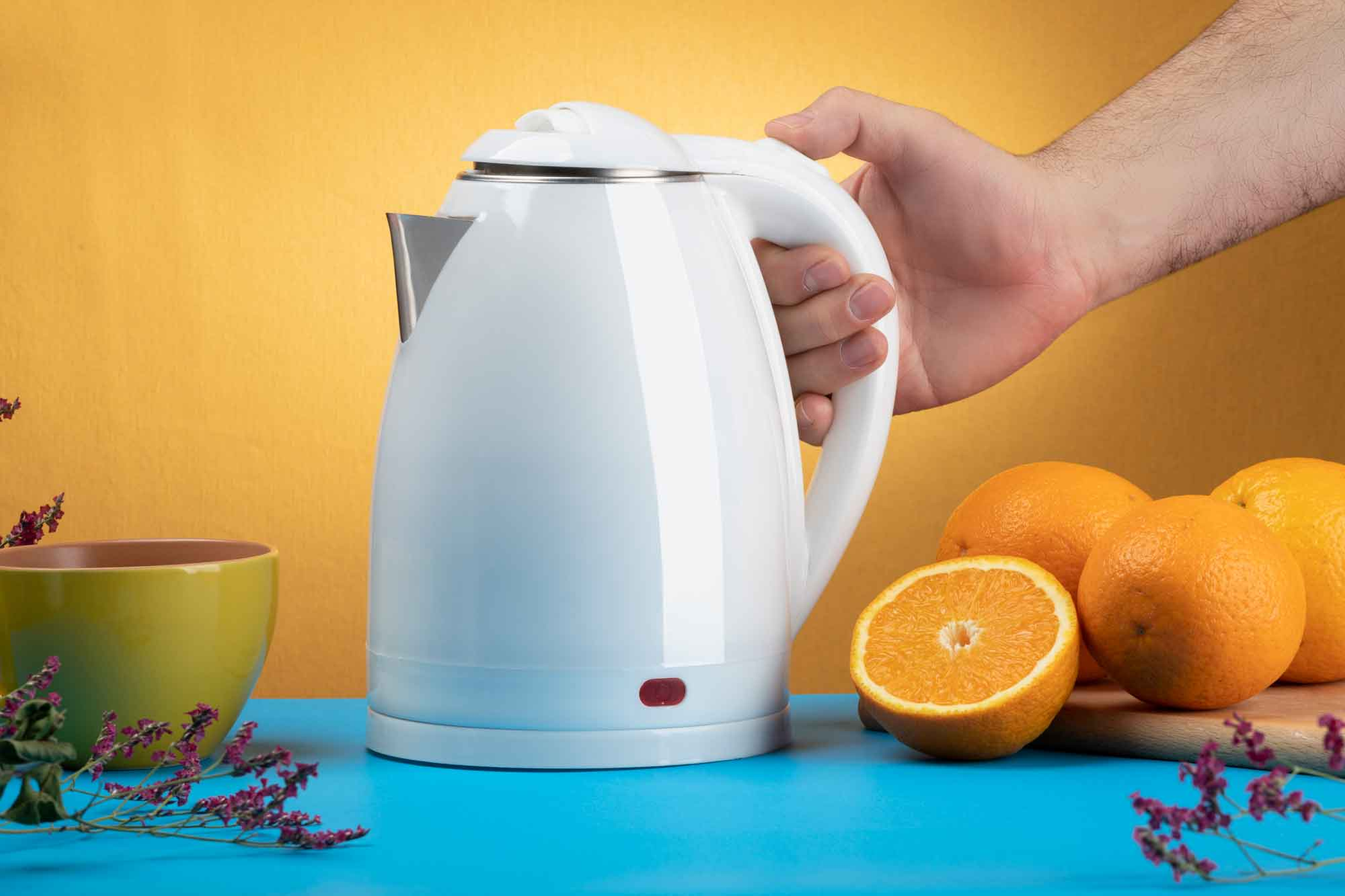 Una tetera eléctrica blanca con mano, naranjas, una taza y flores en la escena.