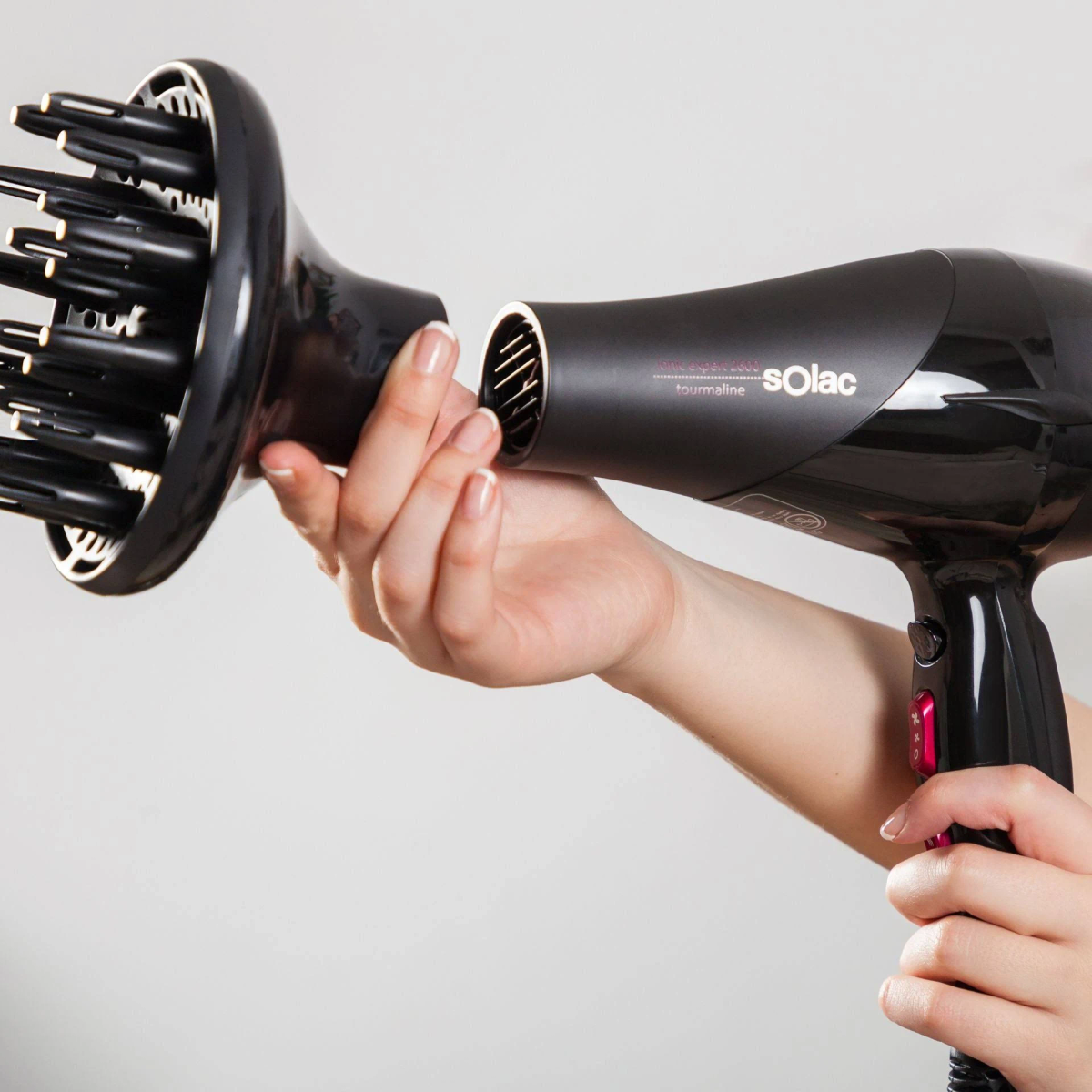 Ręce trzymające czarną suszarkę do włosów, mocujące dyfuzor. Białe tło.