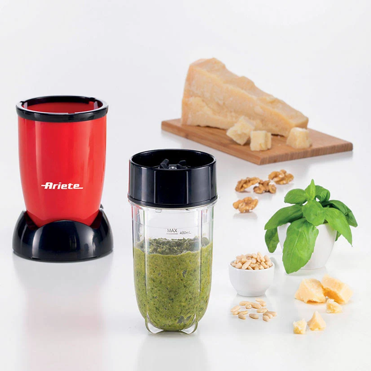 Licuadora con pesto, queso, nueces y albahaca. Licuadora roja a la izquierda, ingredientes dispuestos alrededor.