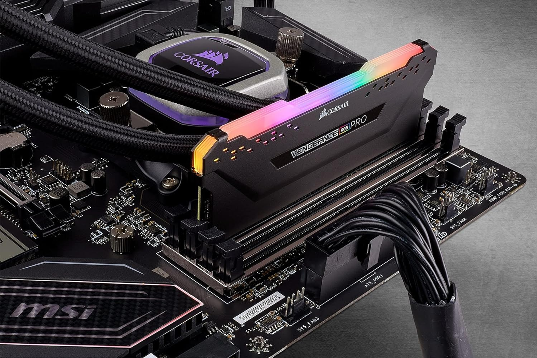 RAM-stick op moederbord, zwart met CORSAIR-label, RGB-verlichting, zwarte kabels. Zilveren koeler.