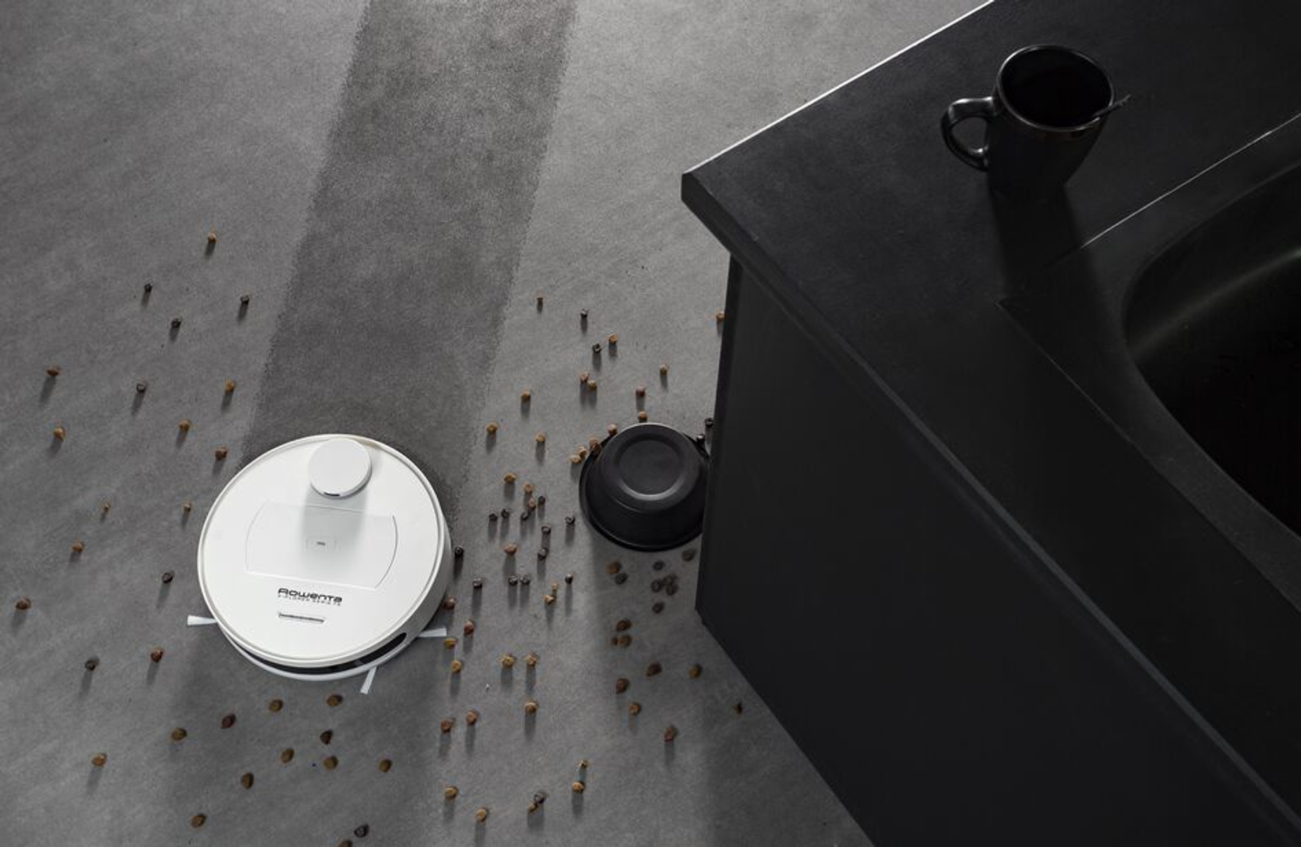 Robot sprzątający na podłodze, czyszczący rozrzucone brązowe granulki. Czarna szafka po prawej. Szara podłoga.